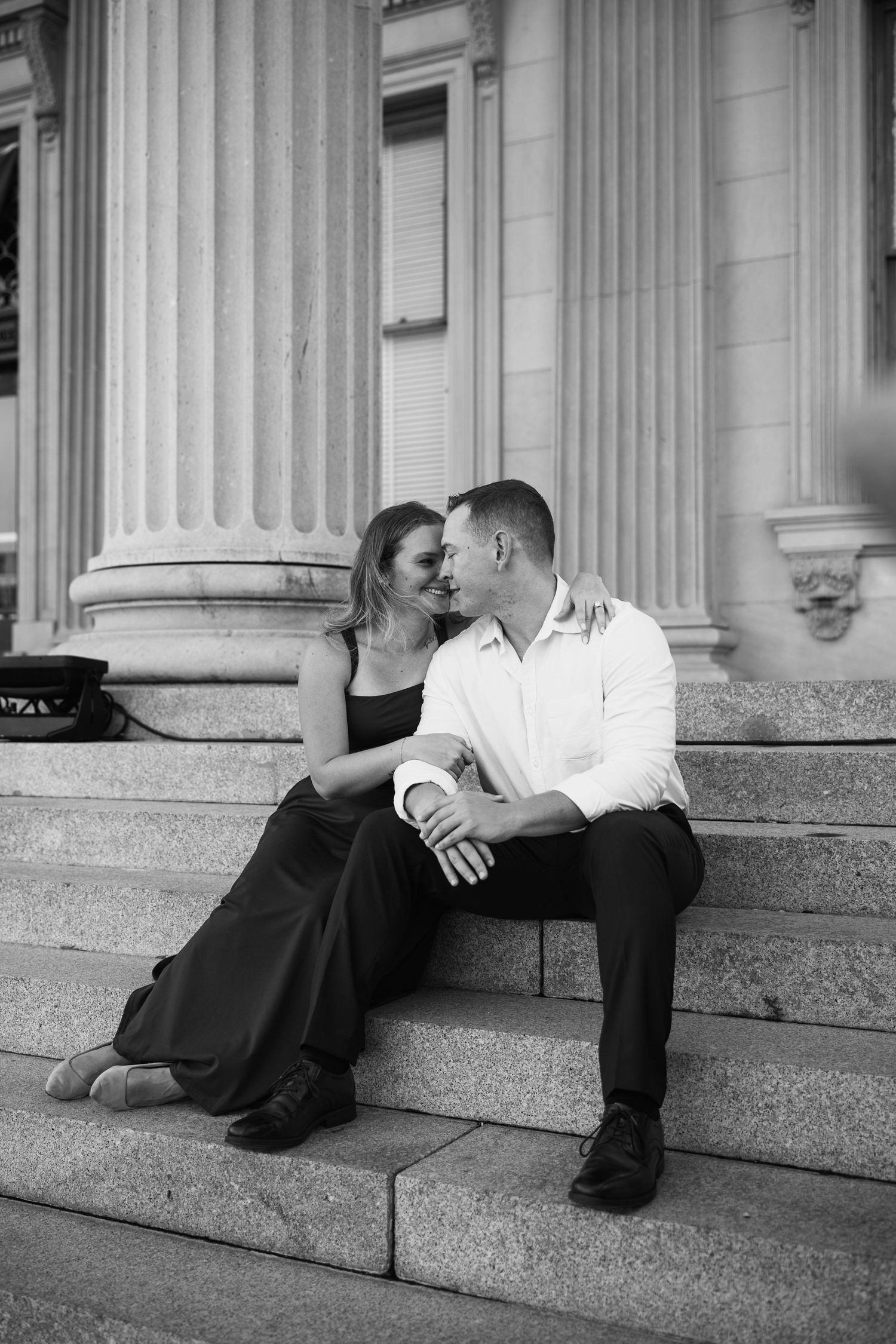 Black and white engagement photo of a couple sitting close on grand stone steps in front of tall columns.