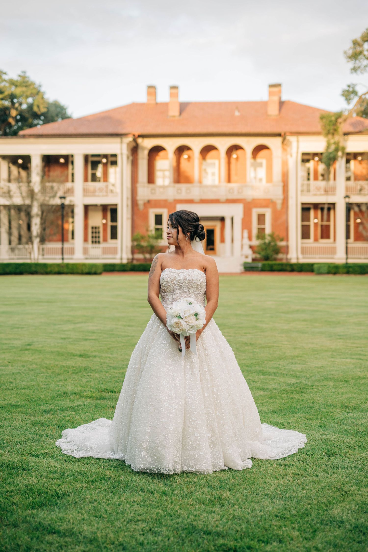 Bride in a strapless sparkling ball gown holding a white bouquet on a green lawn before a red brick Southern mansion.