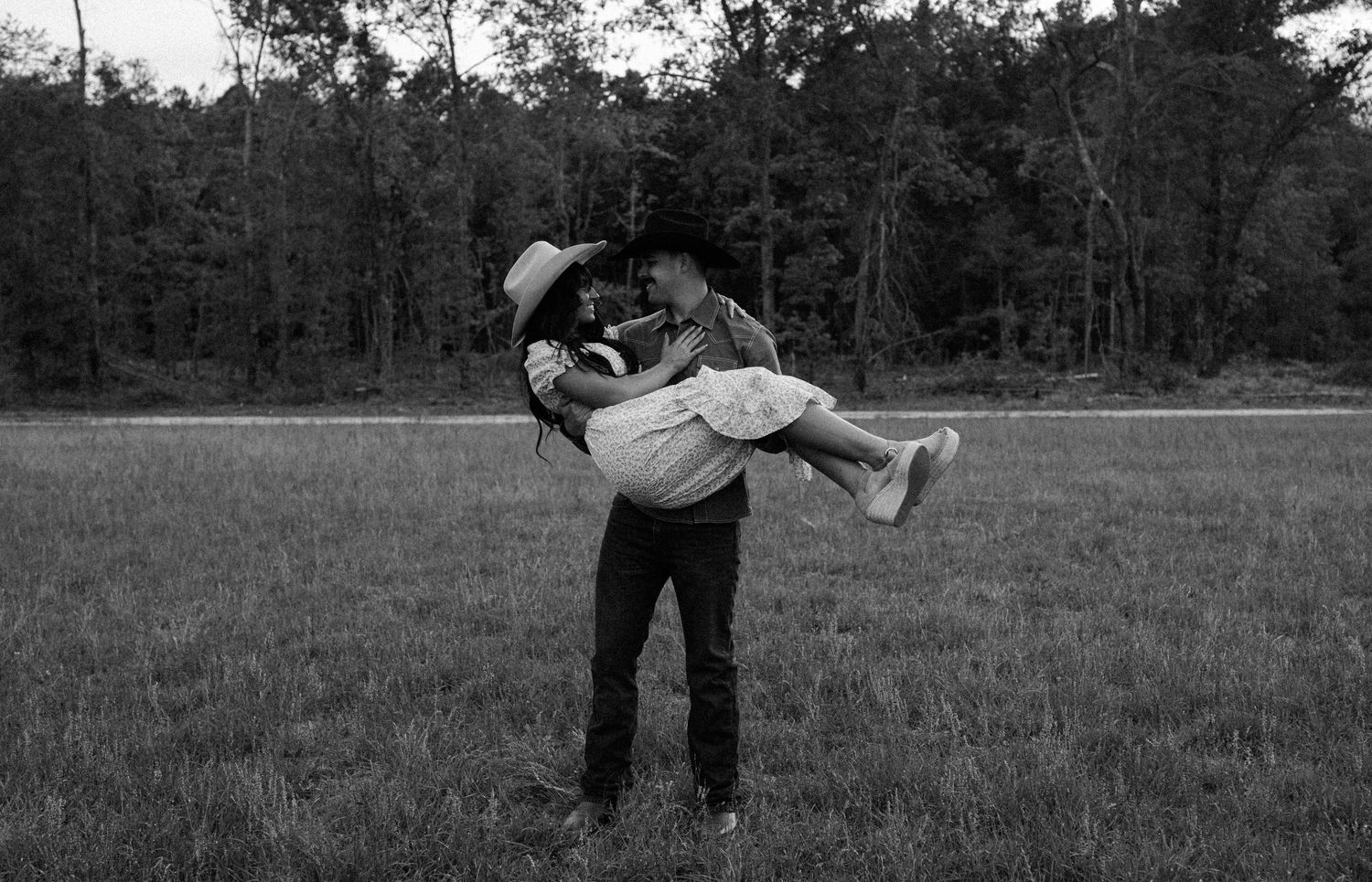 Black and white image of a man in a cowboy hat lifting his partner in his arms in an open grassy field.