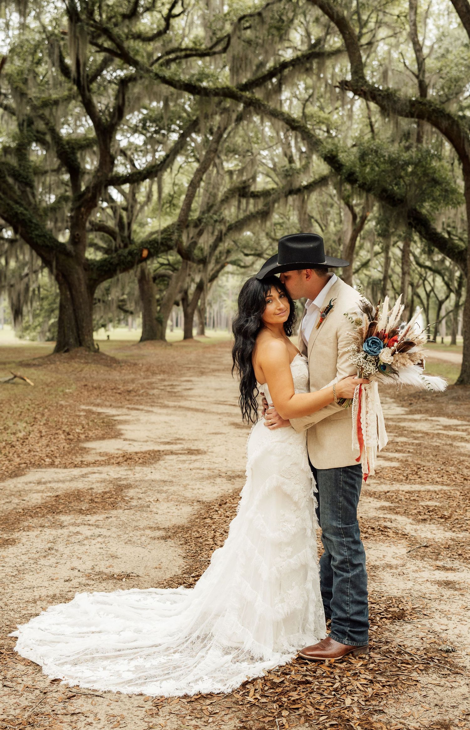 Full-length wedding portrait of a bride with a long lace train and a cowboy-hatted groom embracing on a path under Spanish moss oaks.