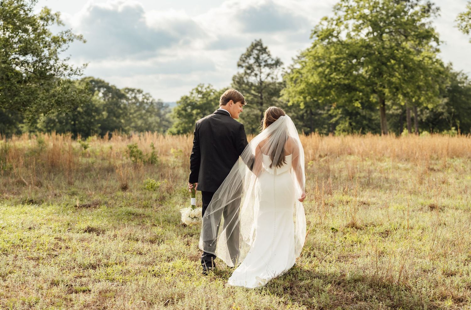 Newlyweds walk hand in hand through a grassy meadow at golden hour, the bride's sheer veil trailing behind her.