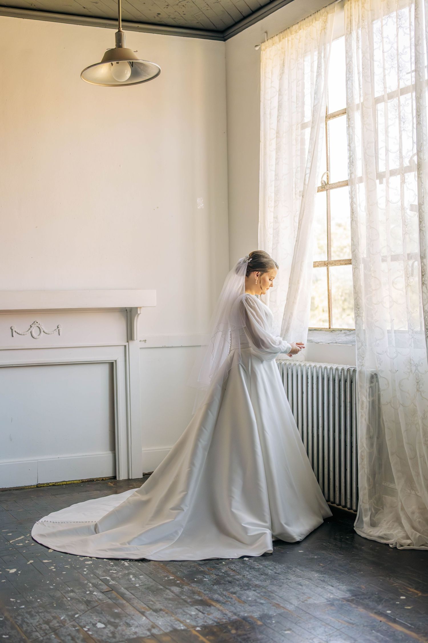 Bride in an elegant satin wedding gown with a long train and veil stands by a lace-curtained window in a bright vintage room.
