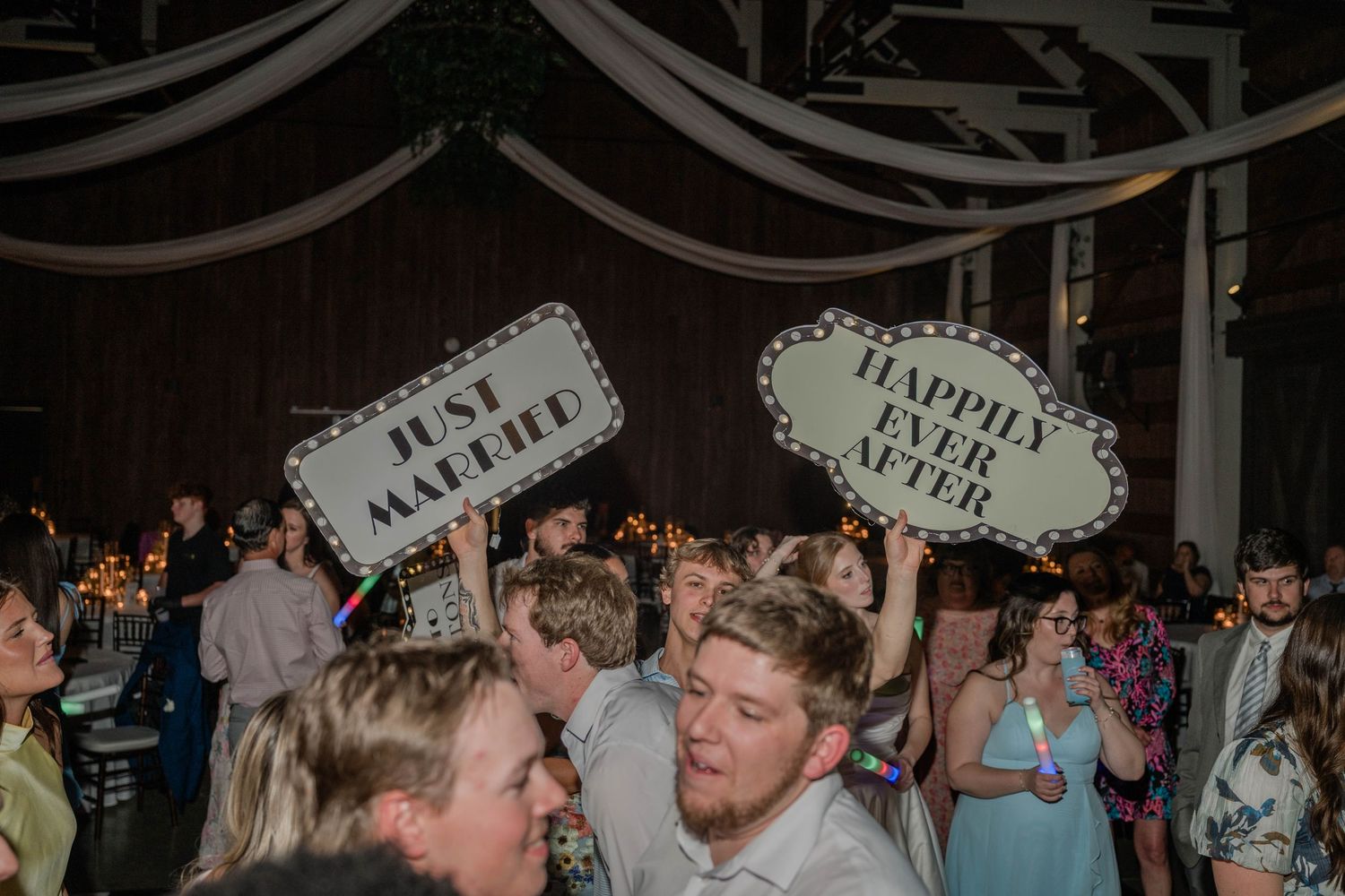 Wedding guests celebrate on the dance floor holding Just Married and Happy Ever After signs under draped reception ceiling fabric.