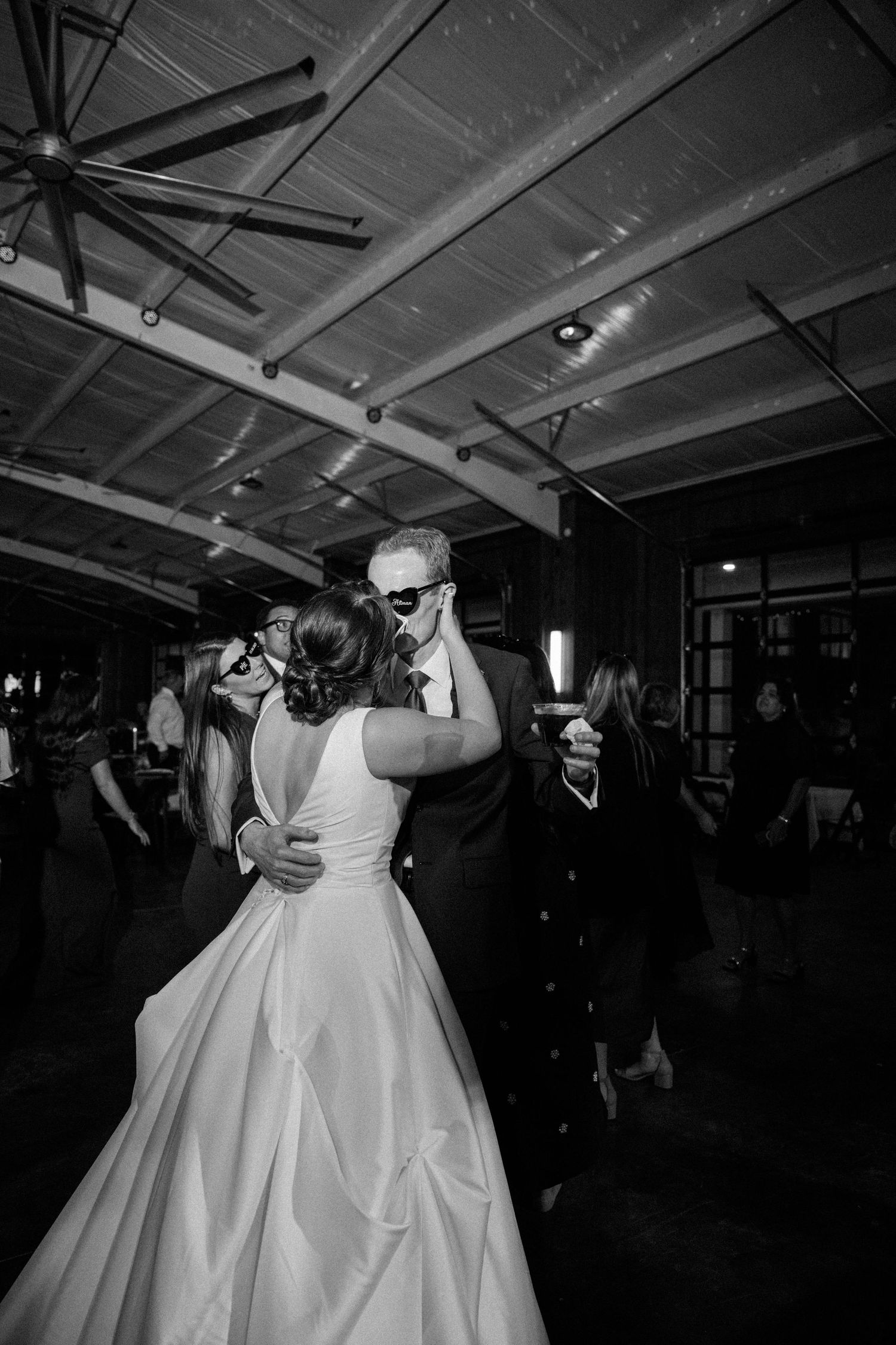 A bride and groom embrace on the dance floor in a black and white photo while guests dance beneath a rustic ceiling fan.