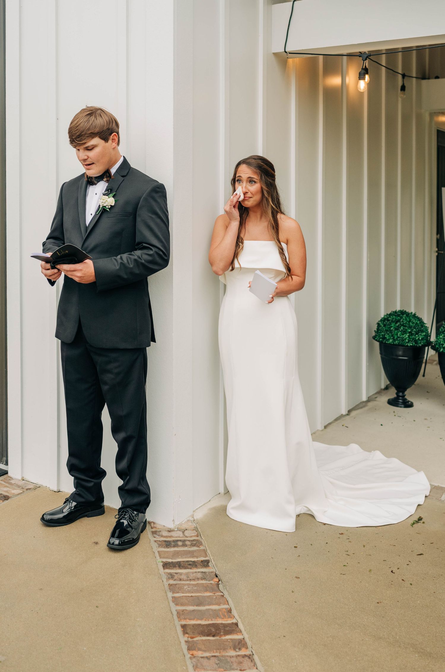 A groom in a black tuxedo reads a letter while a bride in a white gown wipes away tears during a private first look on a porch.