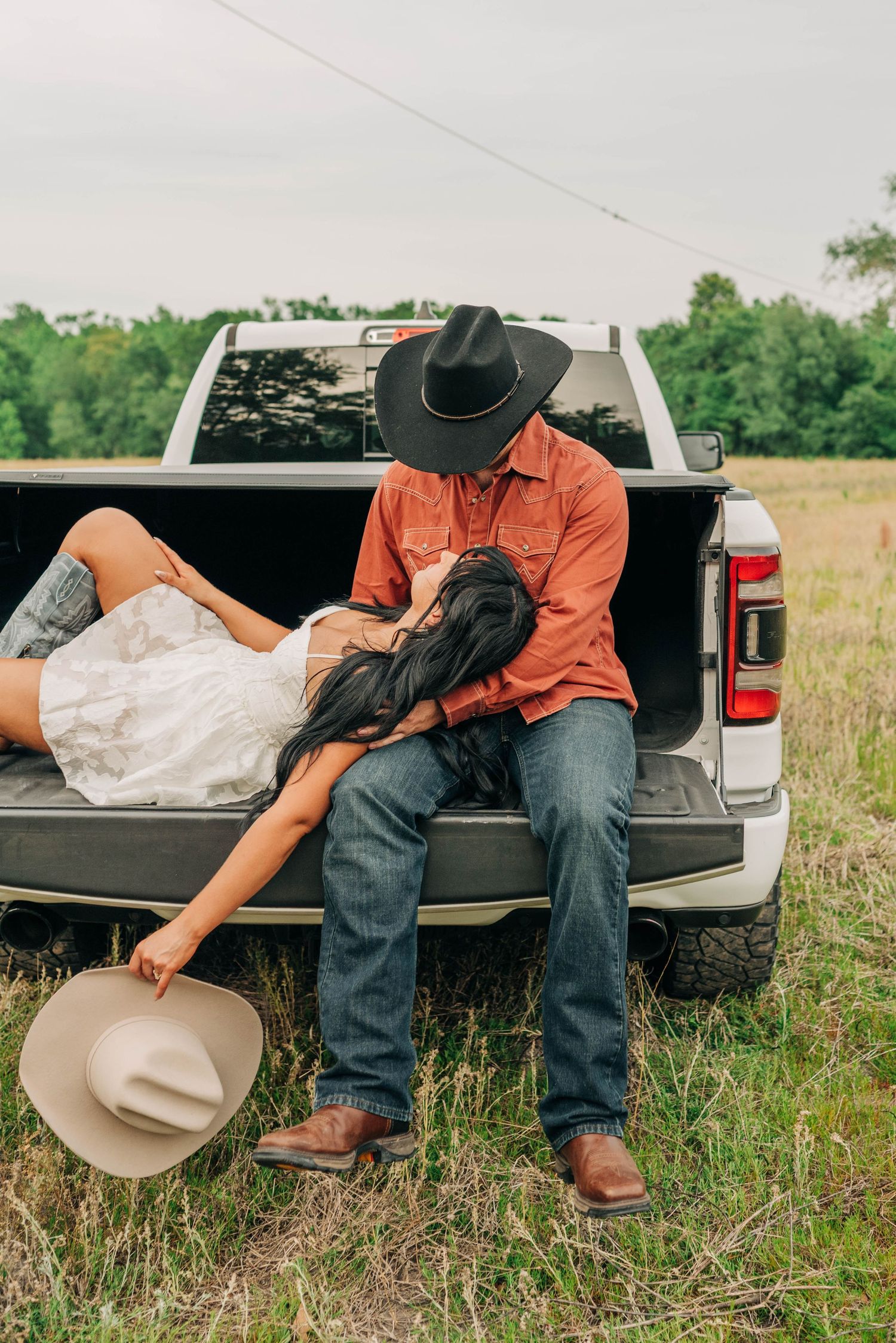 Woman lies in the lap of a man in a cowboy hat and orange shirt on a white truck tailgate in a country field.