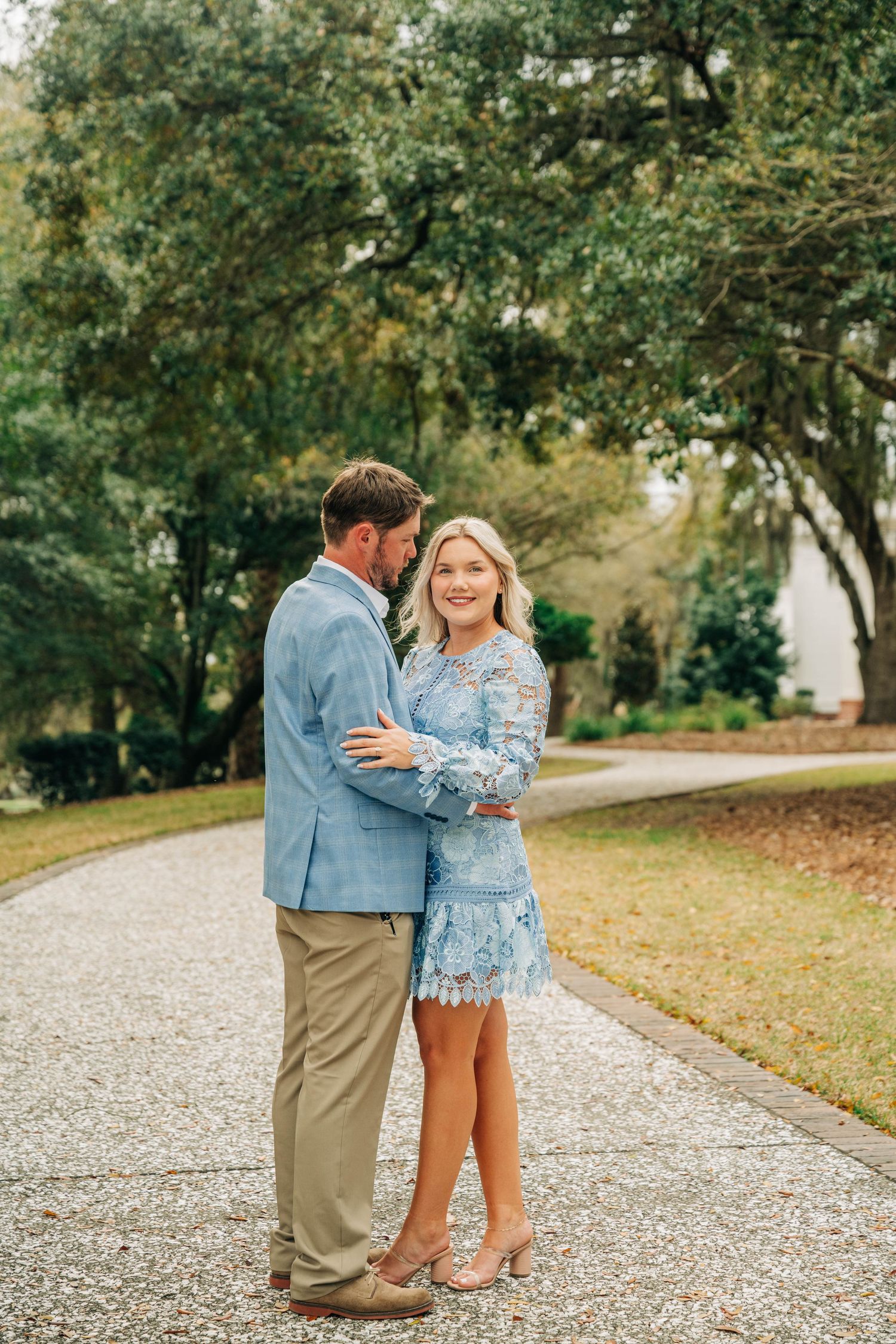 Smiling couple in blue outfits embracing on a gravel path lined with trees during an engagement photo session.