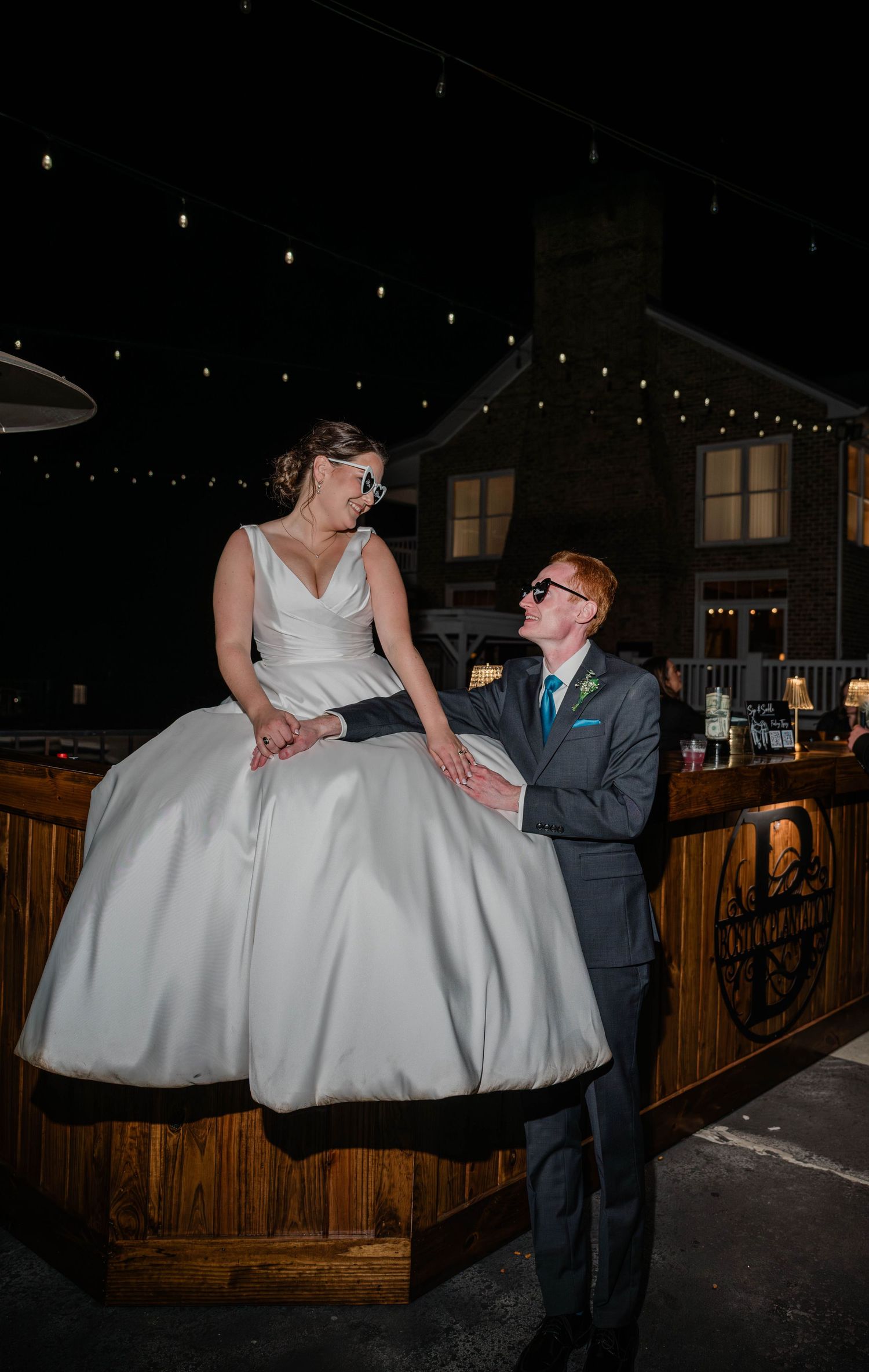 Bride in a satin ball gown sits on a rustic wooden bar as the groom gazes up at her under evening string lights.