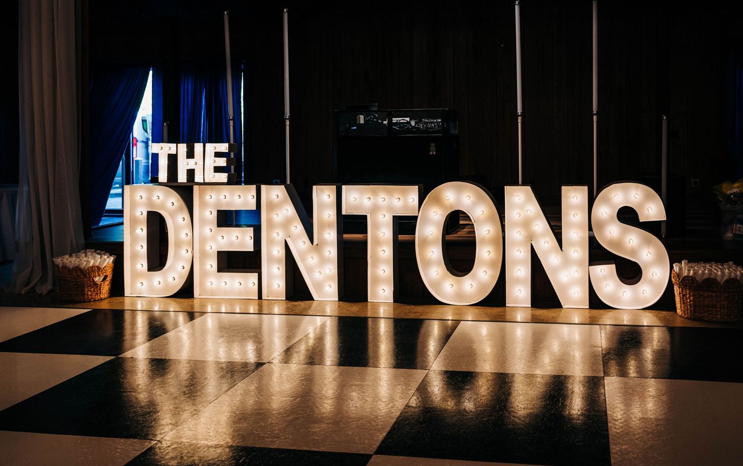 Illuminated marquee letters spelling The Dentons stand on a black and white checkered dance floor at a wedding reception.