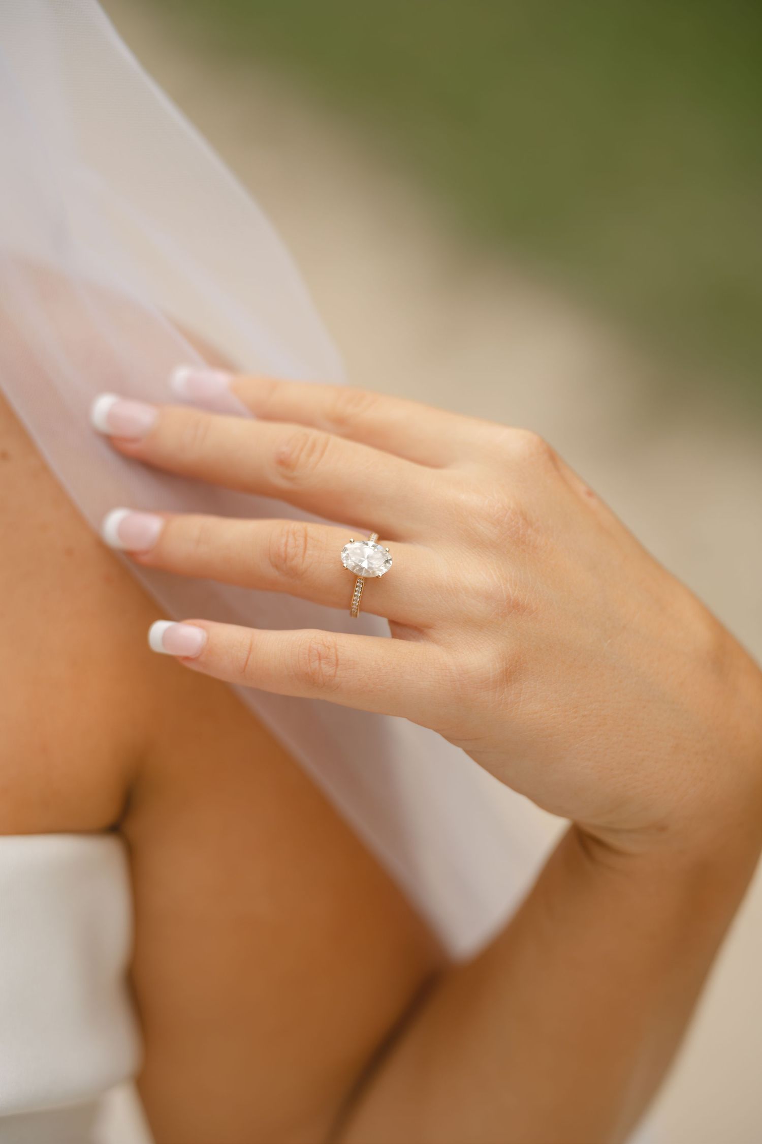Close-up of a bride's hand with an oval diamond engagement ring and pale manicure resting on her veil.