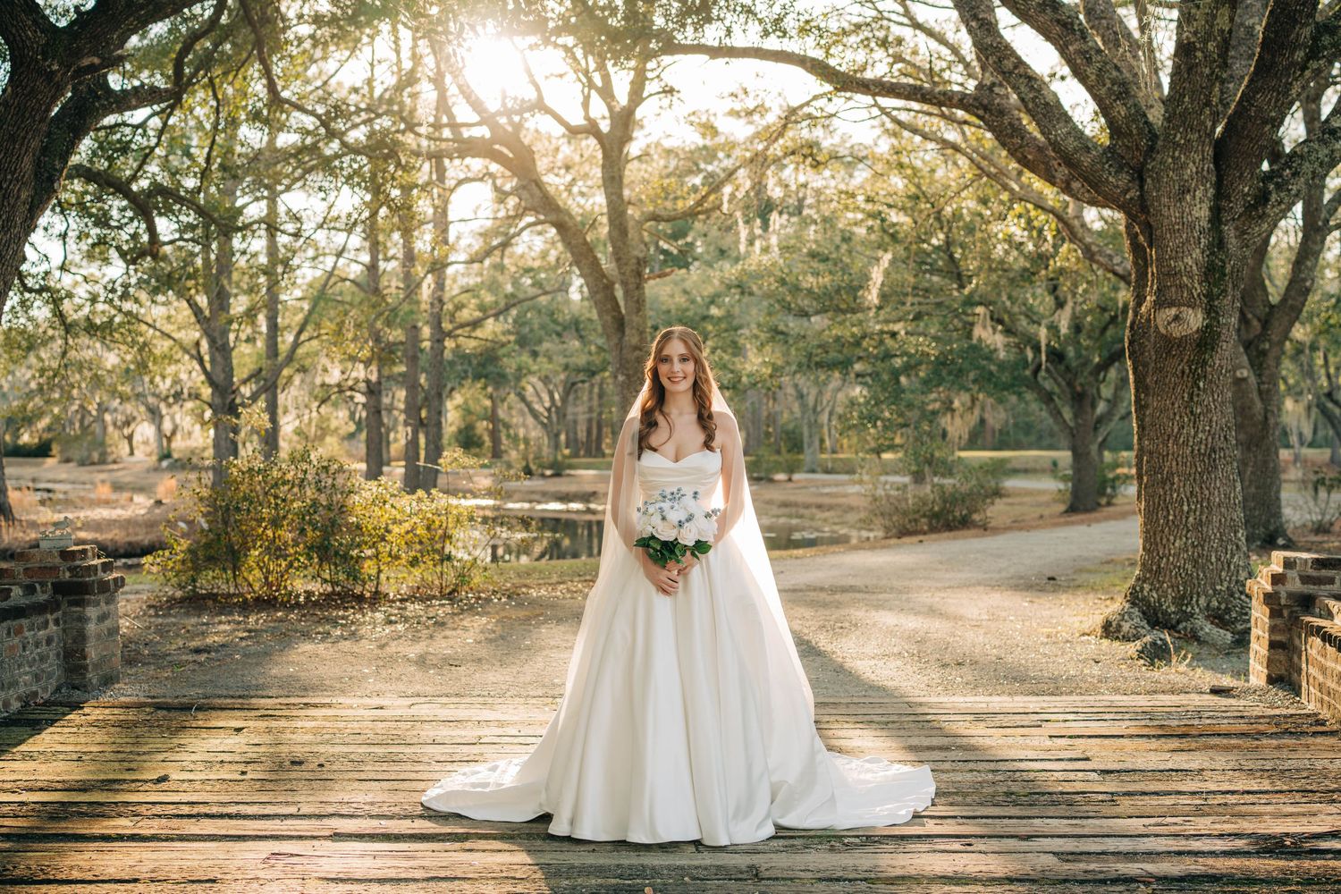 Bride in a strapless white gown with a long veil holding a bouquet on a wooden deck beneath sunlit oak trees.