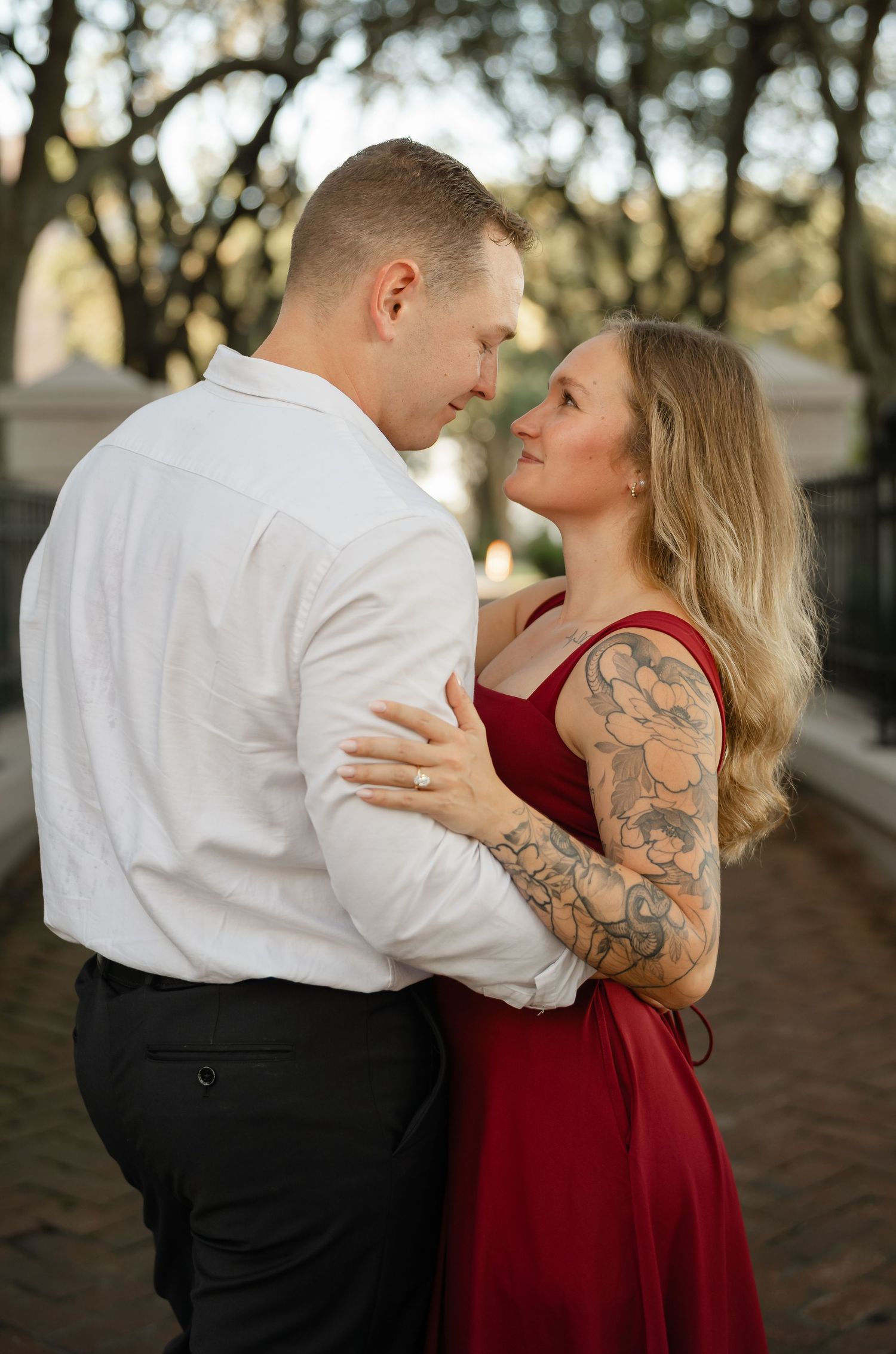 Engaged couple in a romantic embrace on a brick path, the woman in a red dress with a floral tattoo and the man in a white shirt.