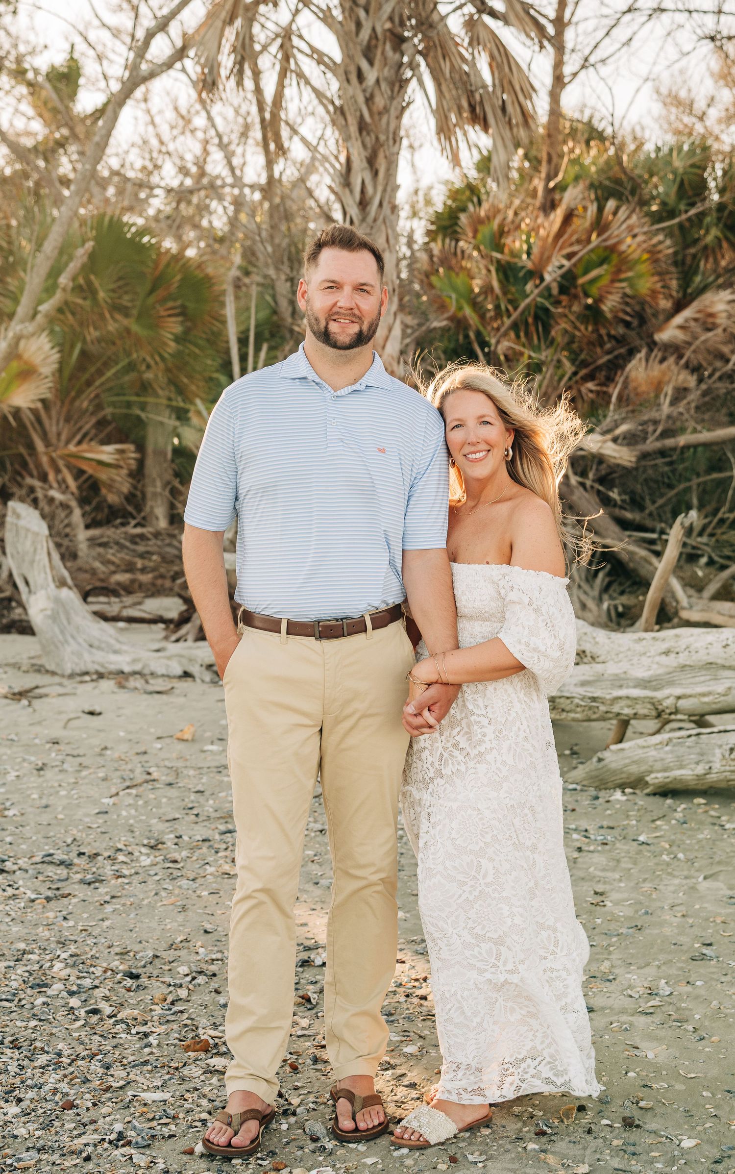 Smiling couple in light summer outfits embrace on a sandy beach surrounded by palm trees during an engagement photo session.