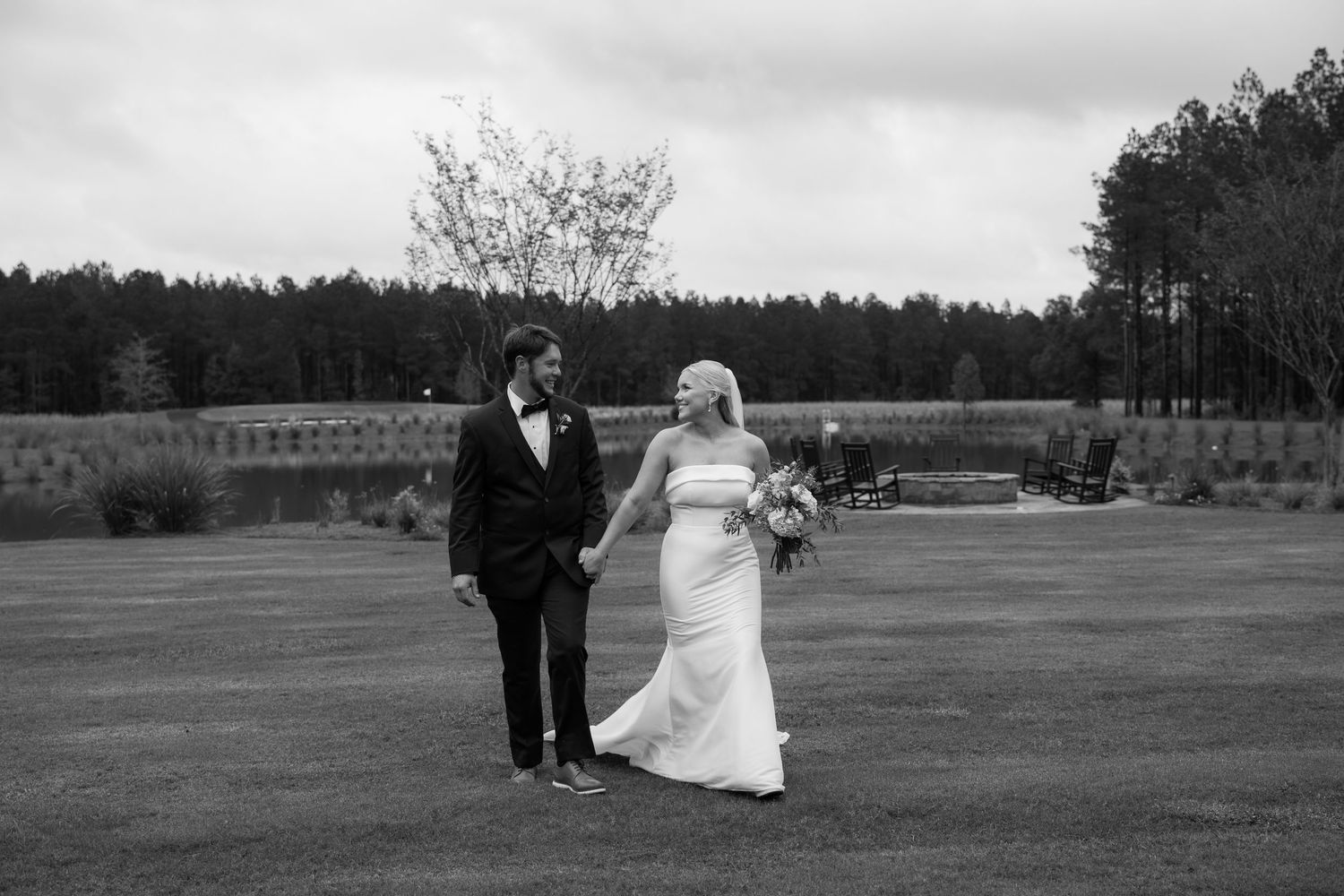 Black and white photo of a bride holding a bouquet and walking hand in hand with her groom across an open lawn.