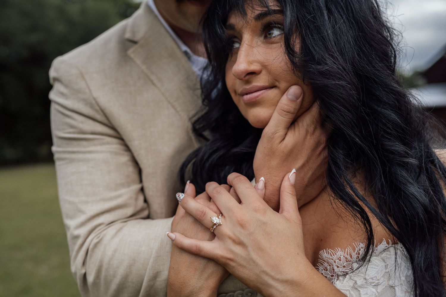 Close-up of a bride with long dark hair and a diamond ring gazing upward as her groom in a tan blazer embraces her from behind.