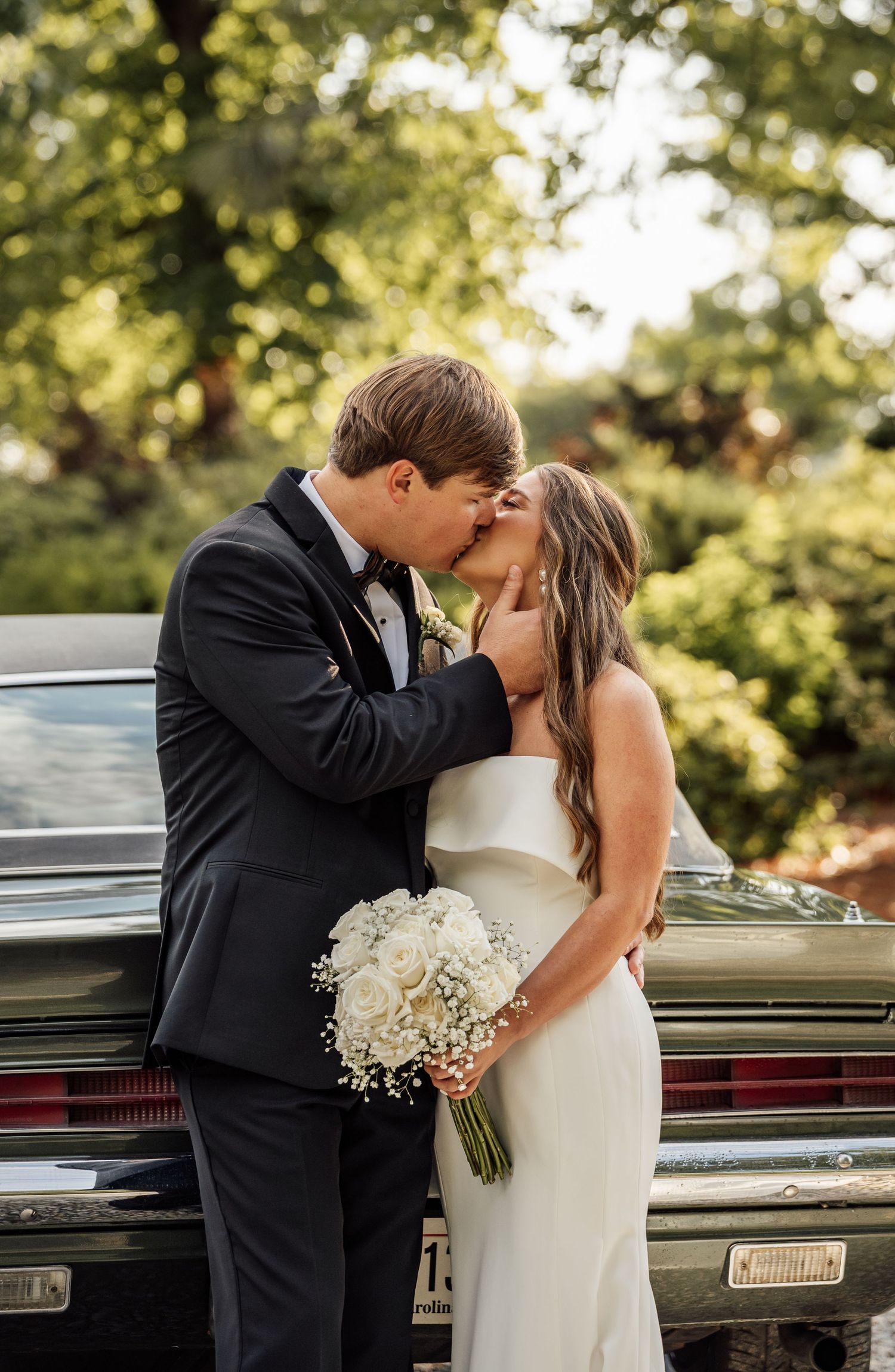 Close-up of a bride and groom kissing in front of a vintage car, the bride holding a bouquet of white roses.