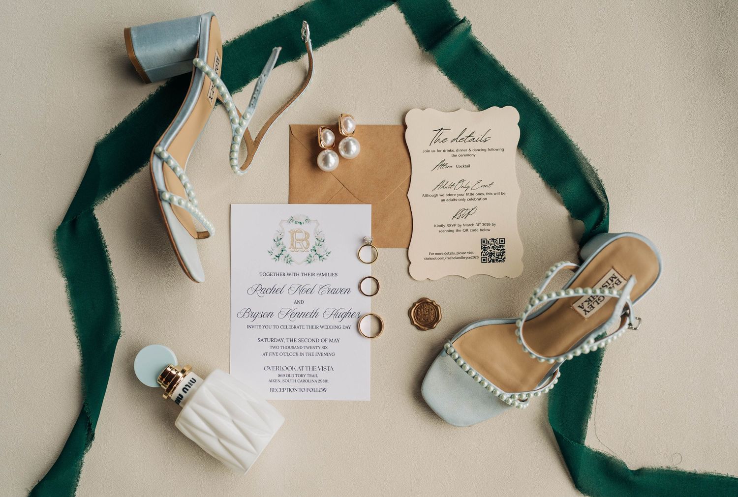 Flat lay of wedding invitation, gold rings, pearl earrings, perfume and pearl-trimmed blue heels styled with an emerald green ribbon.