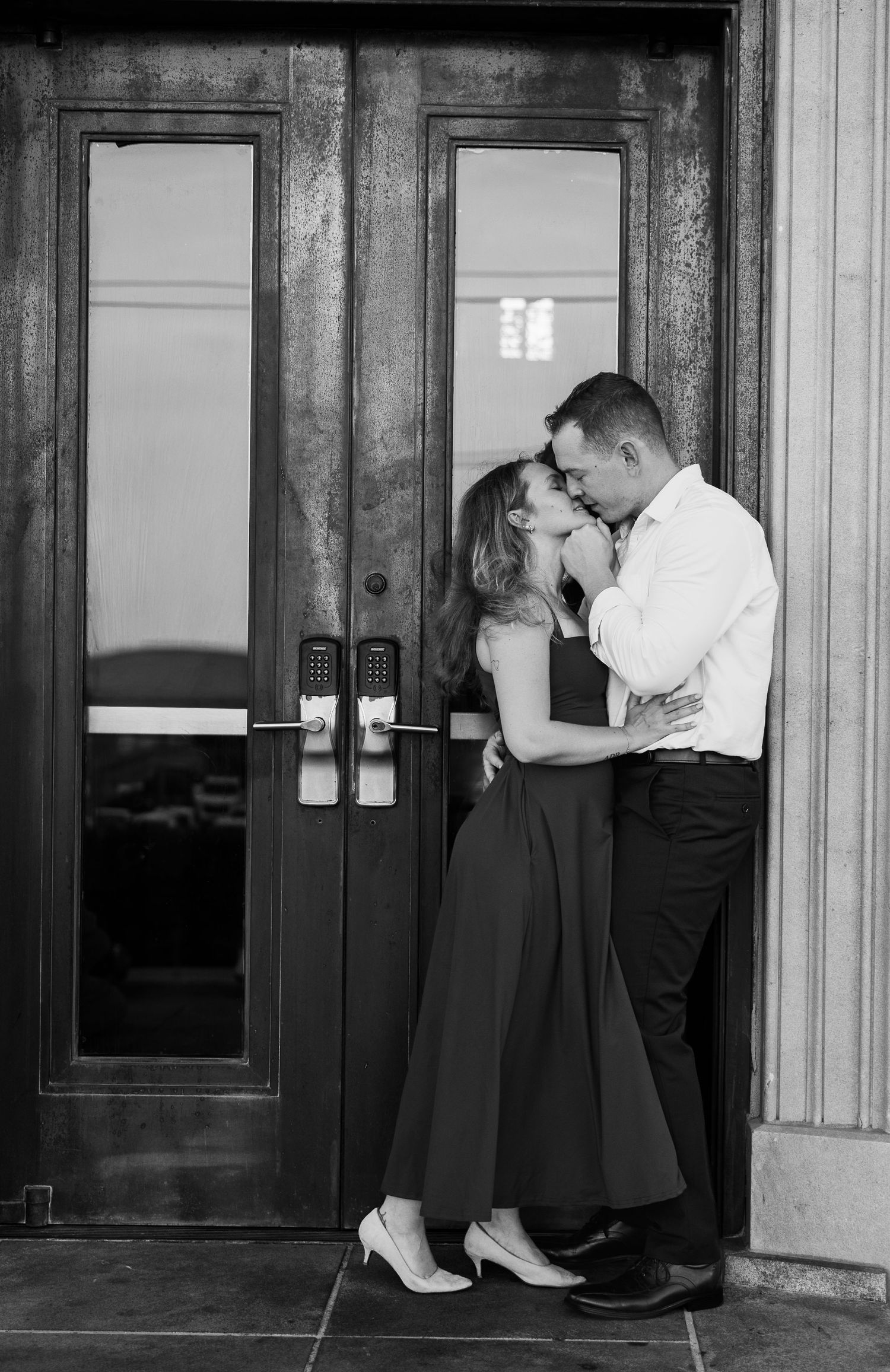 Black and white engagement photo of a couple kissing in front of vintage double doors, the woman in a long dark dress and heels.
