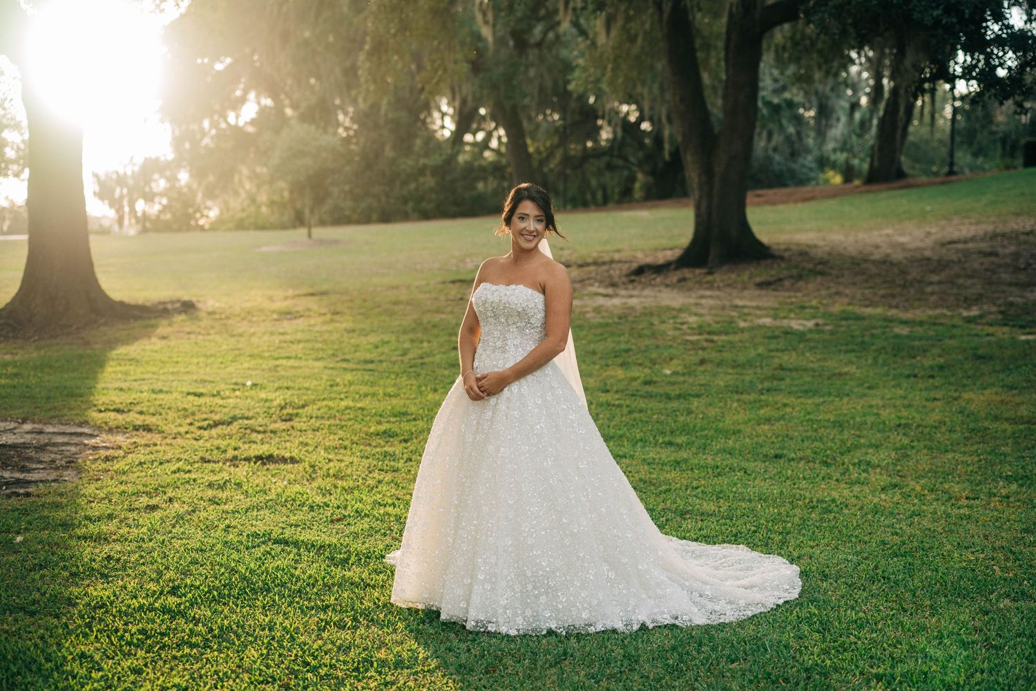 Bride in a tulle ball gown with a lace bodice poses on a sunlit green lawn beneath large trees at golden hour.