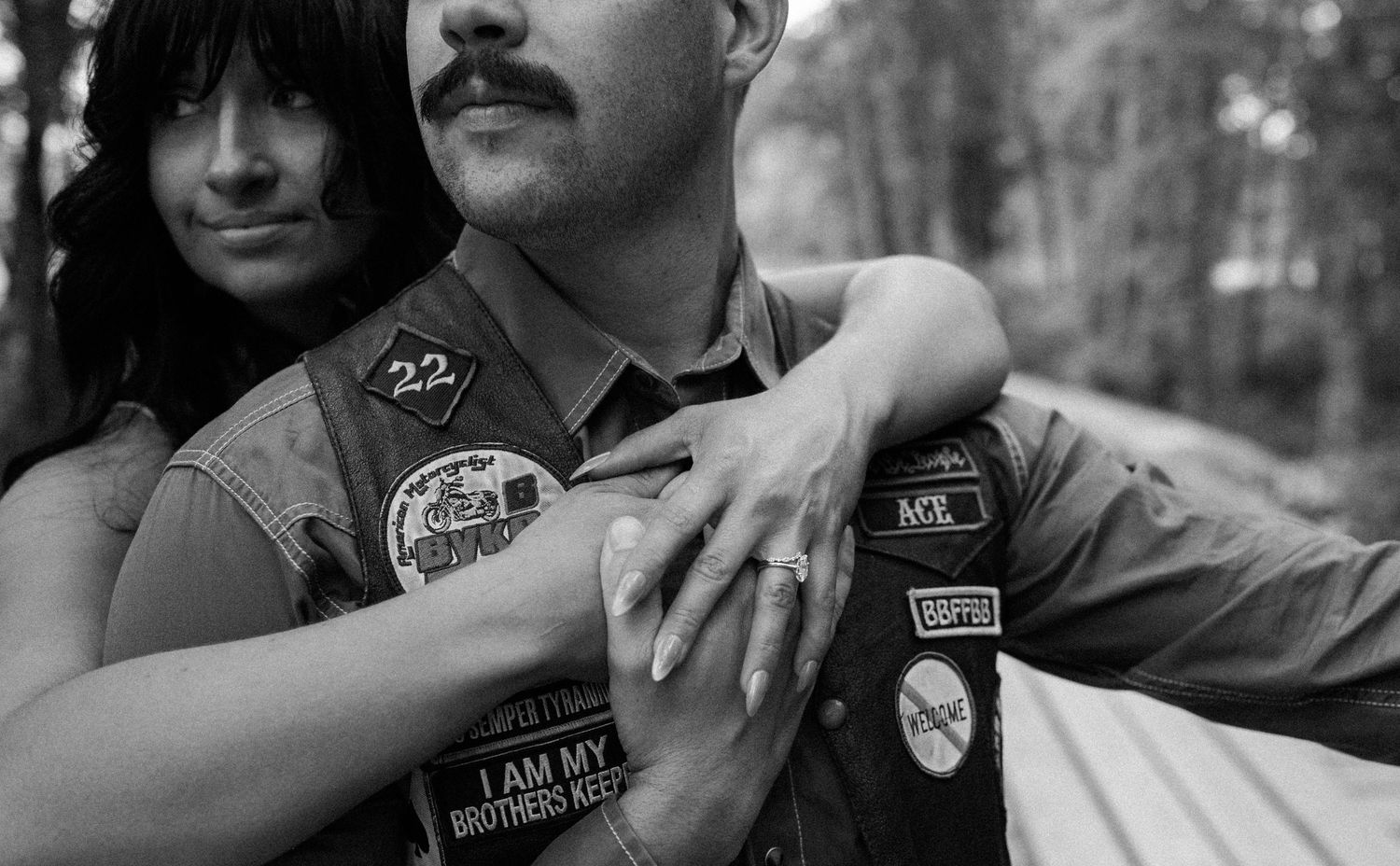 Black and white engagement portrait of a bearded biker in a patched vest embraced from behind by a dark-haired woman.