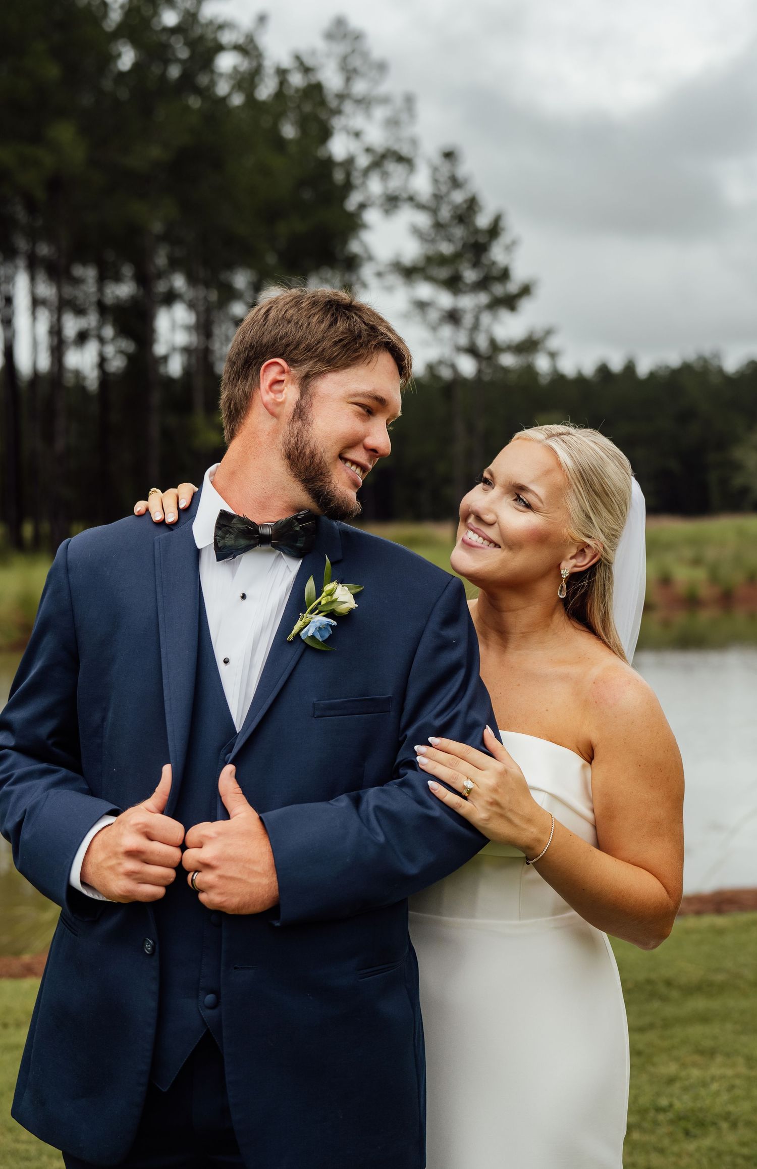 Smiling groom in a navy suit and bow tie embraces his bride in a strapless white gown beside a pond and tall pine trees.