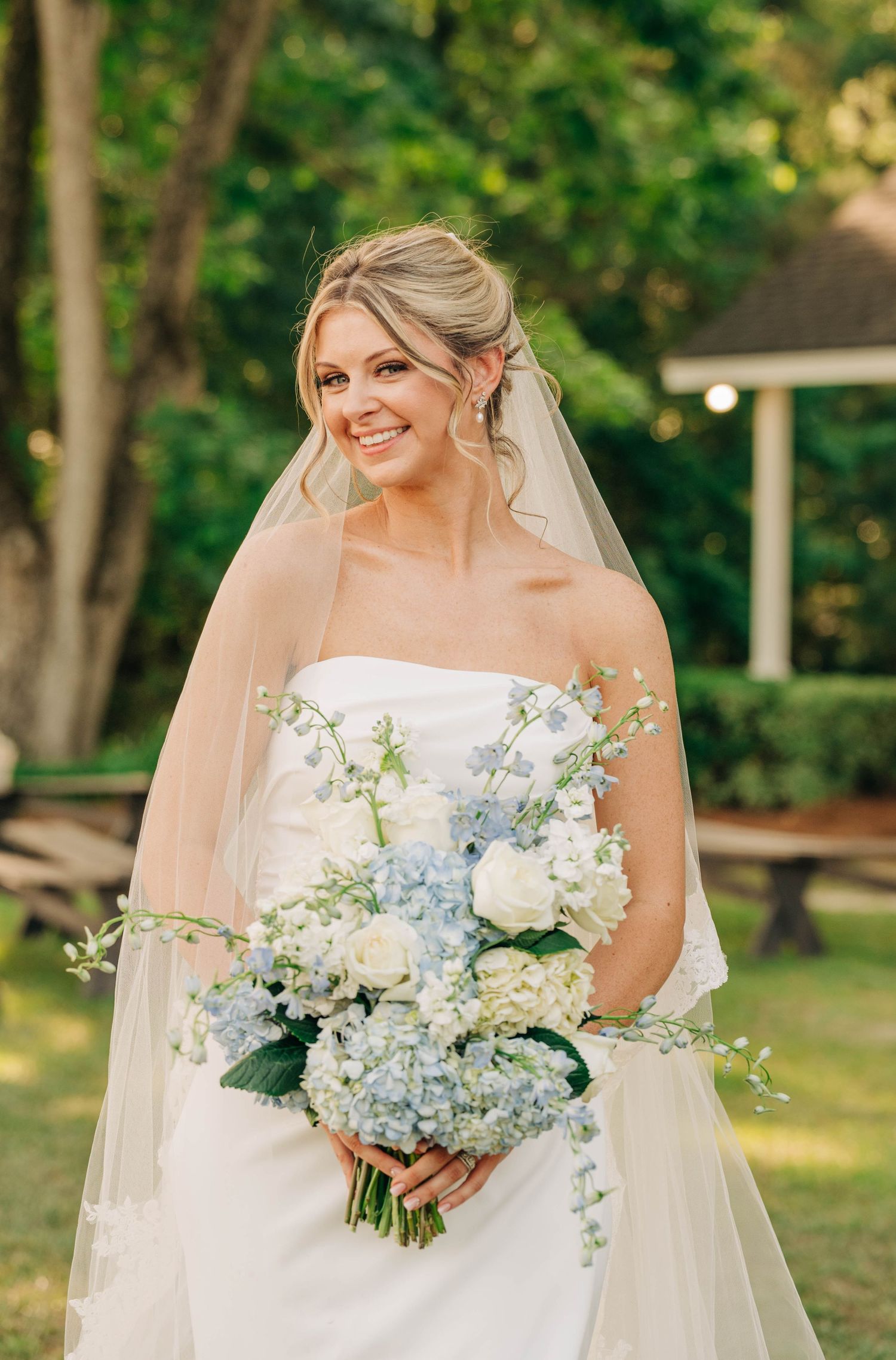 Smiling bride in a strapless gown and sheer veil holds a blue and white hydrangea bouquet in a lush green garden.