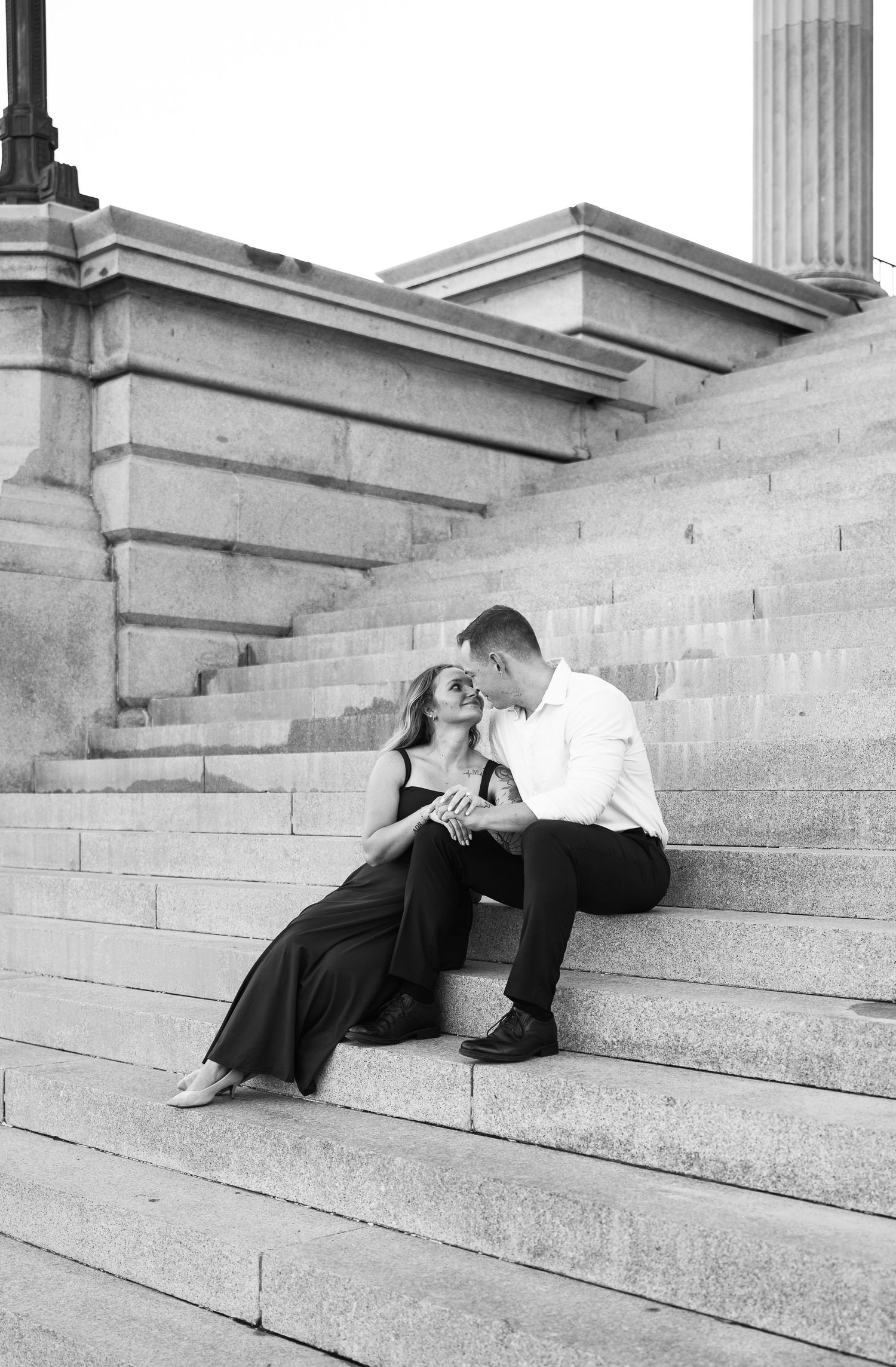 Black and white engagement portrait of a couple sitting on grand stone steps near a column, smiling and holding hands.