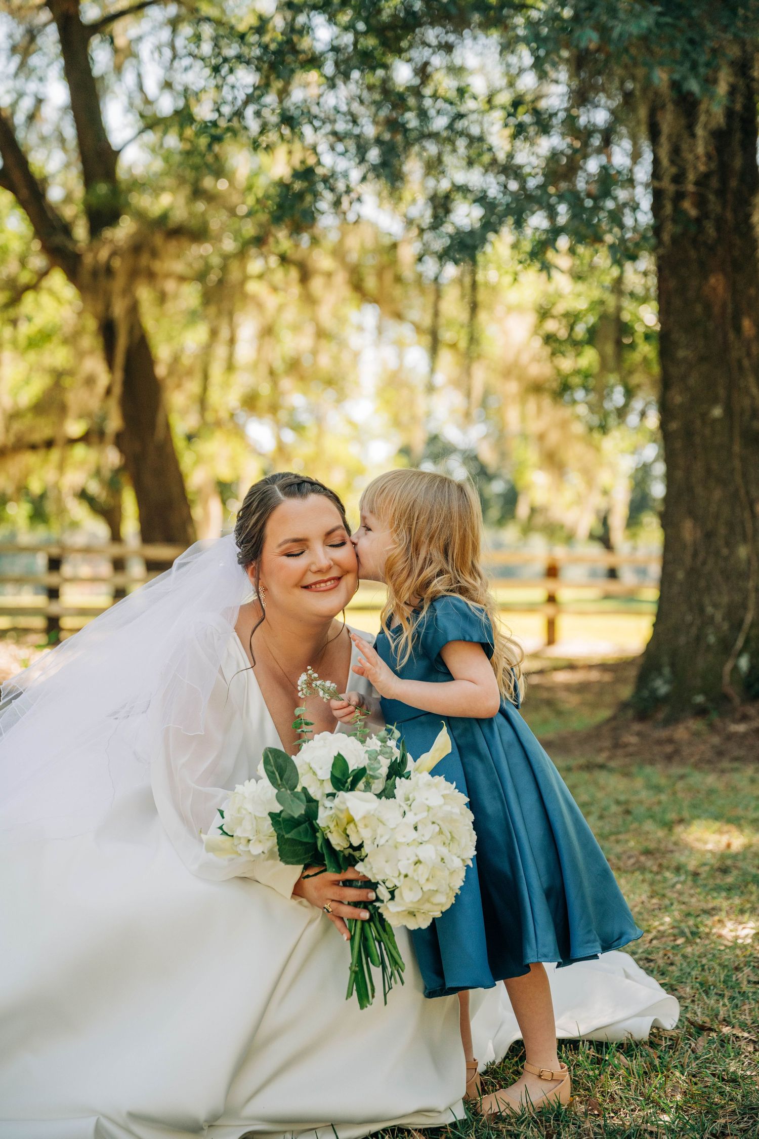 Smiling bride in a white gown holding a white floral bouquet receives a kiss on the cheek from a flower girl in a teal dress.