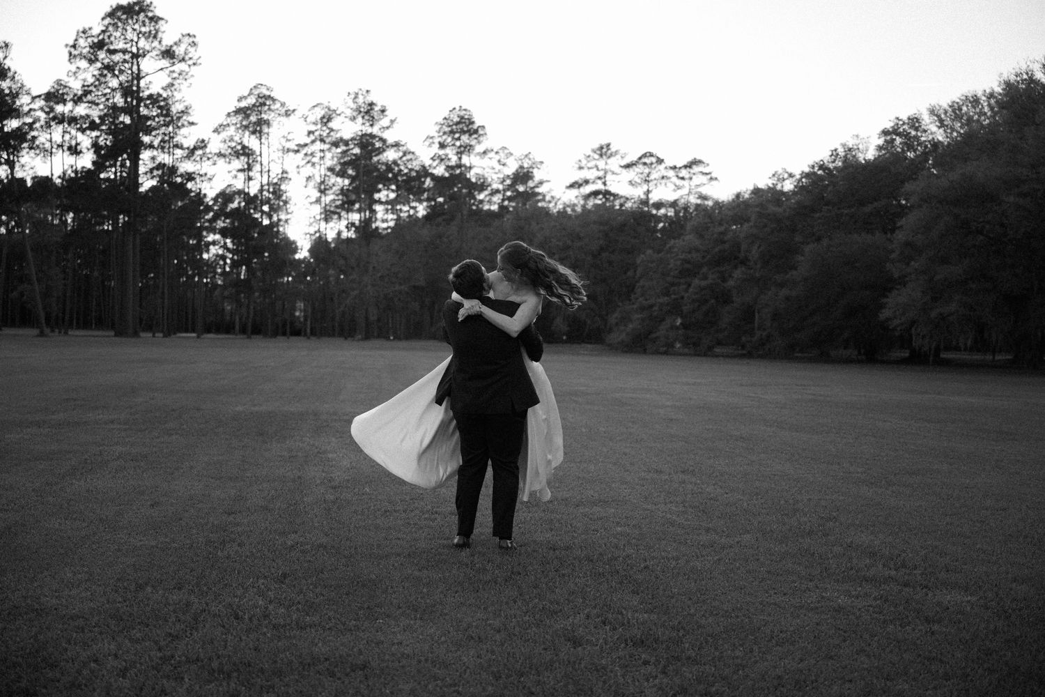 Groom lifts his bride in a flowing white gown across an open grassy field with a tree line behind them in black and white.