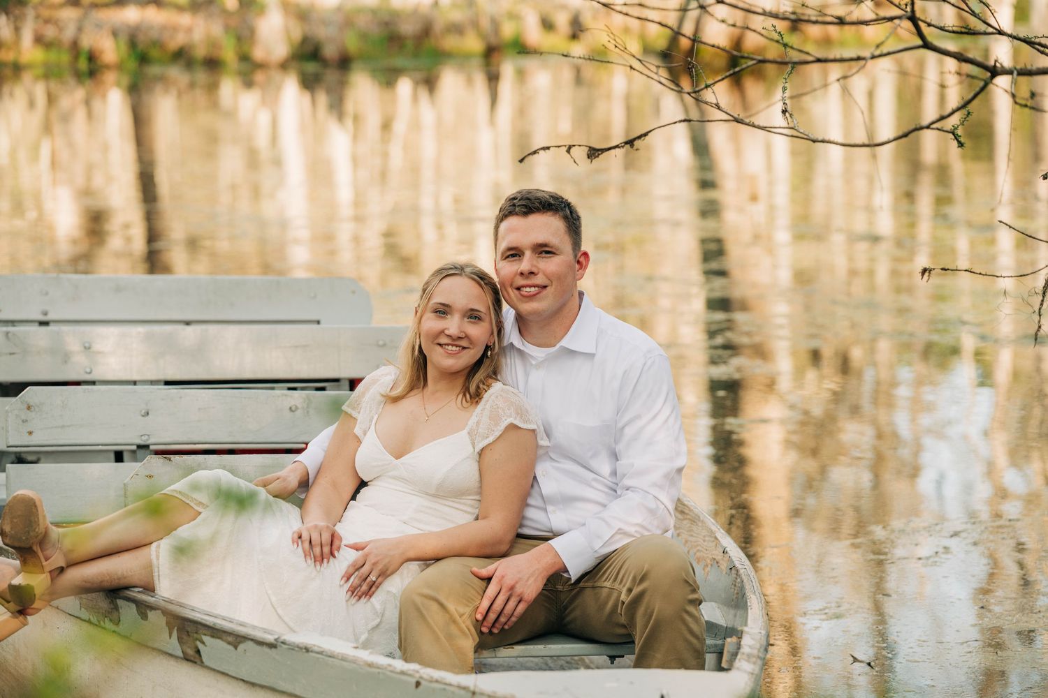 Engaged couple in white sit together in a rowboat on a calm lake reflecting golden autumn trees.