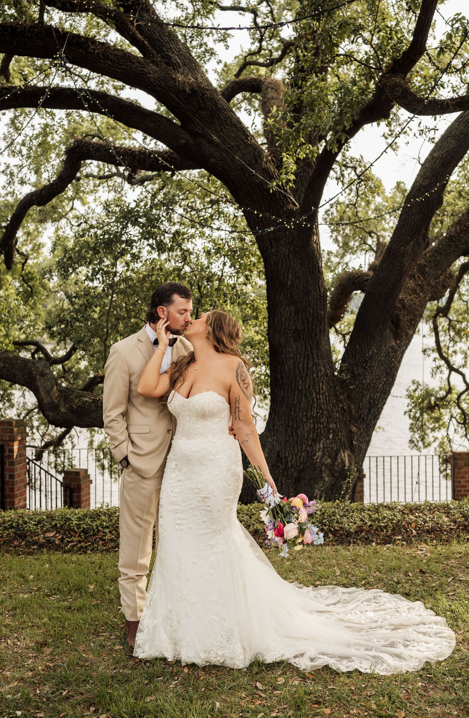 Couple shares a kiss beneath a large oak tree on a lawn, the bride's long lace wedding gown train flowing behind her.