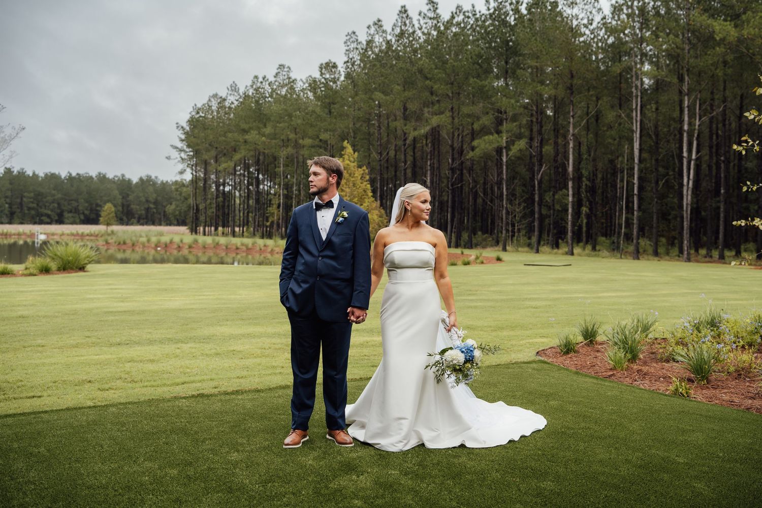 Groom in a navy suit and bride in a fitted white gown hold a bouquet on a green lawn with a pine tree forest behind them.