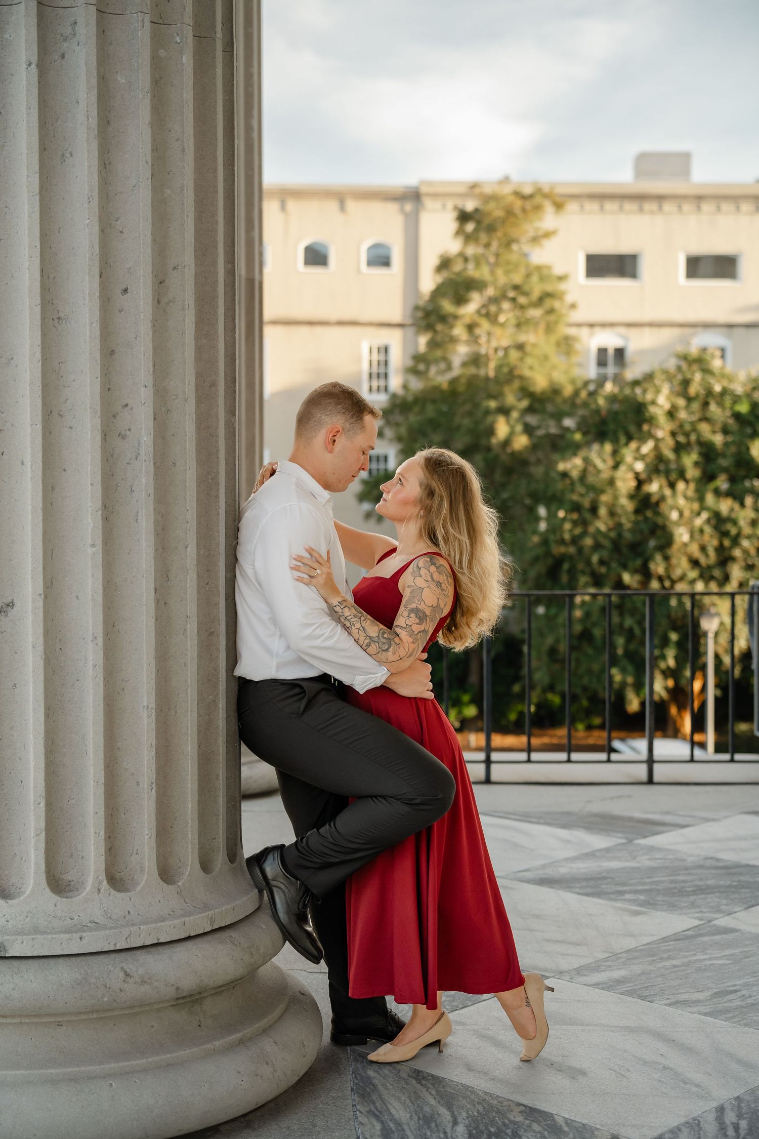 Couple embracing against a tall stone column on a terrace, the woman in a red dress and the man in a white shirt and dark pants.