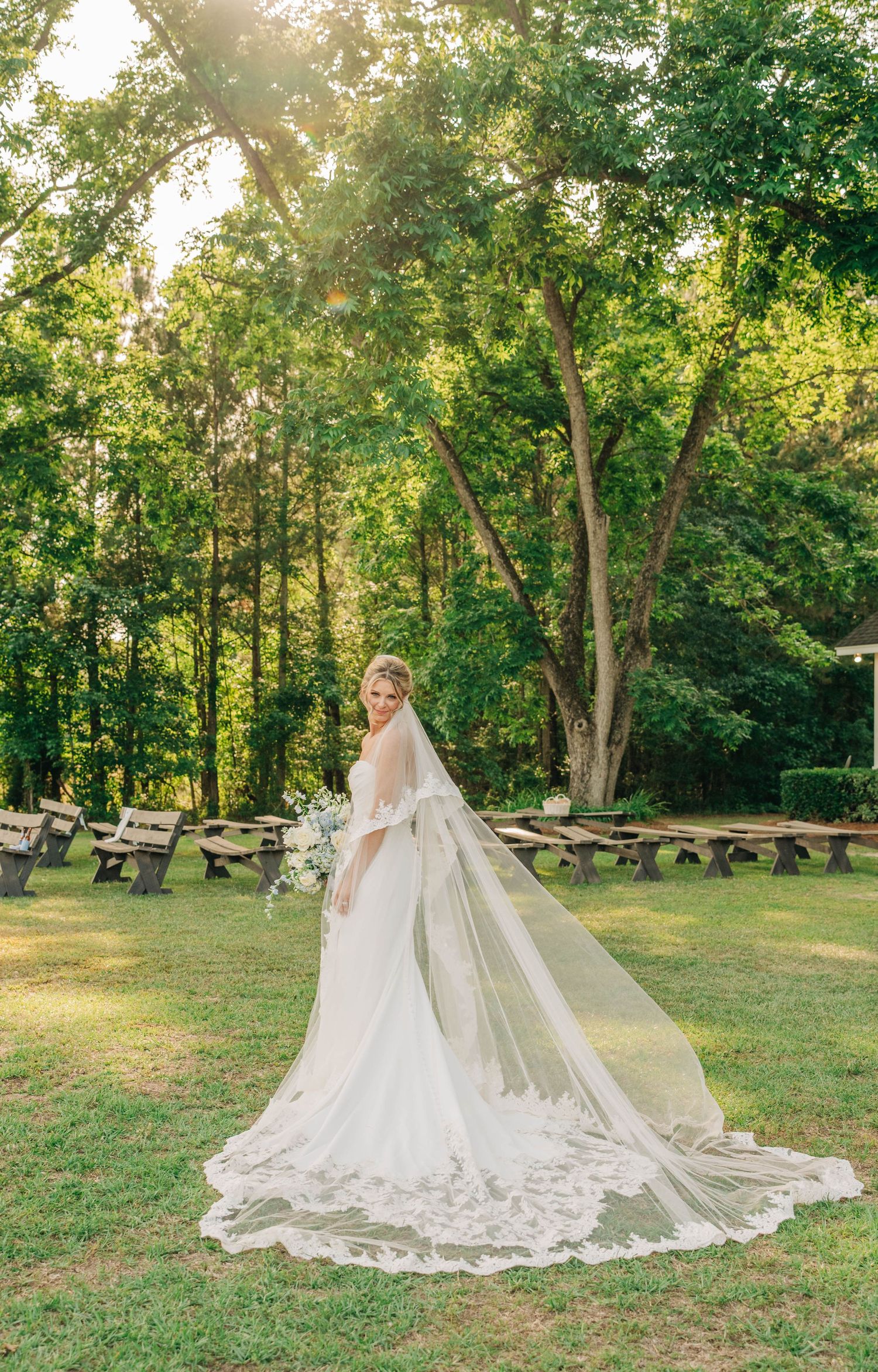 Bride standing on a grassy lawn in a fitted gown with a long lace-edged veil flowing behind her near wooden benches.