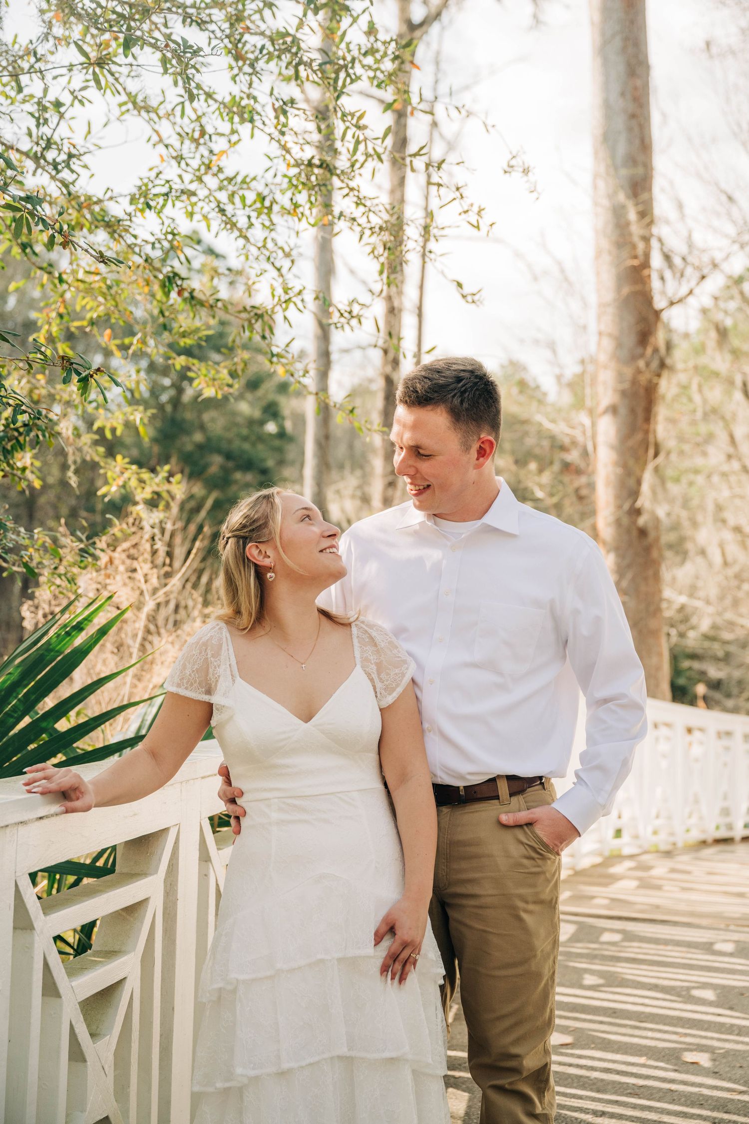 Happy couple smiles at each other on a white wooden bridge surrounded by greenery during an engagement photo session.