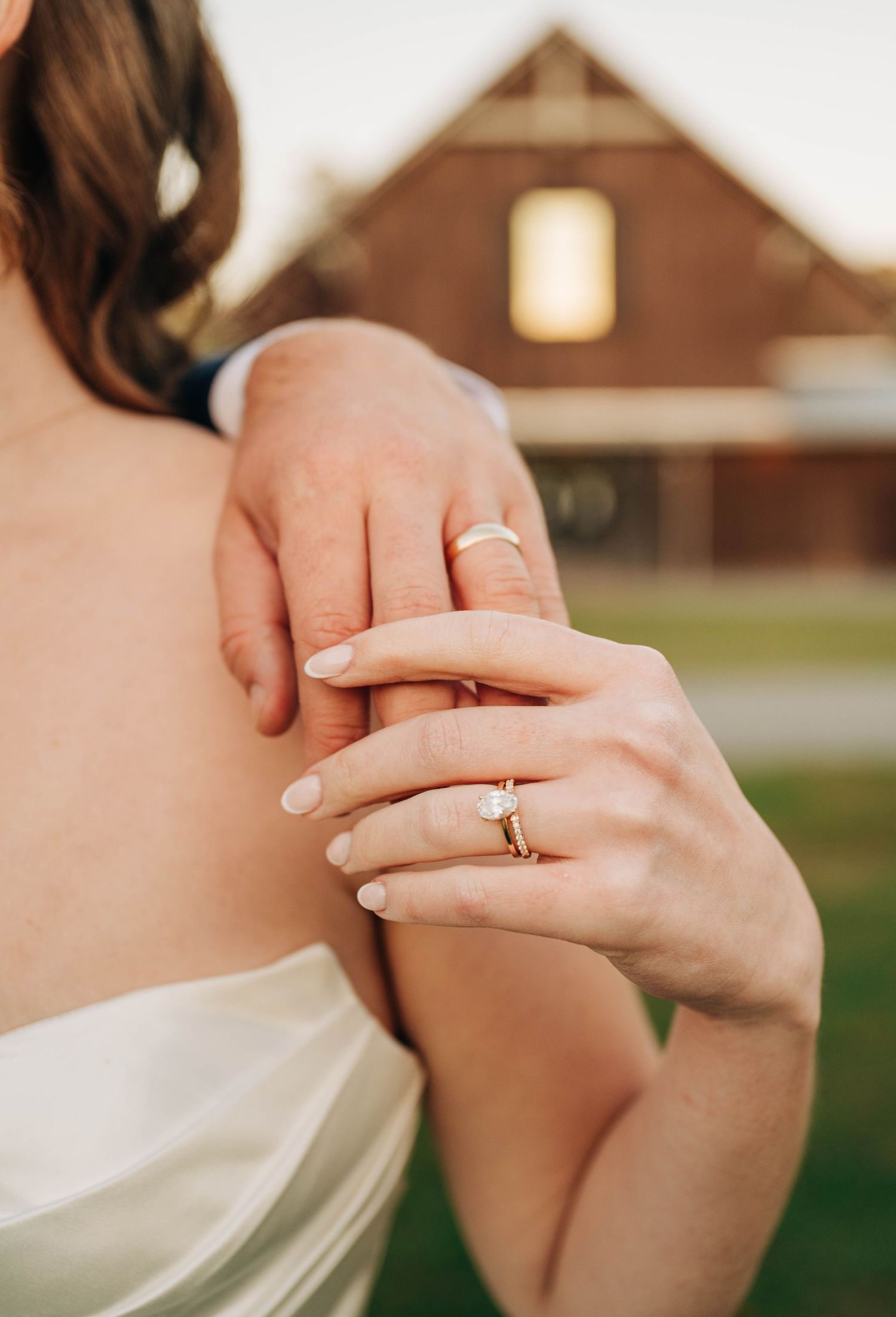 Close-up of the bride and groom's hands wearing wedding rings, with a diamond engagement ring and a rustic barn in the background.