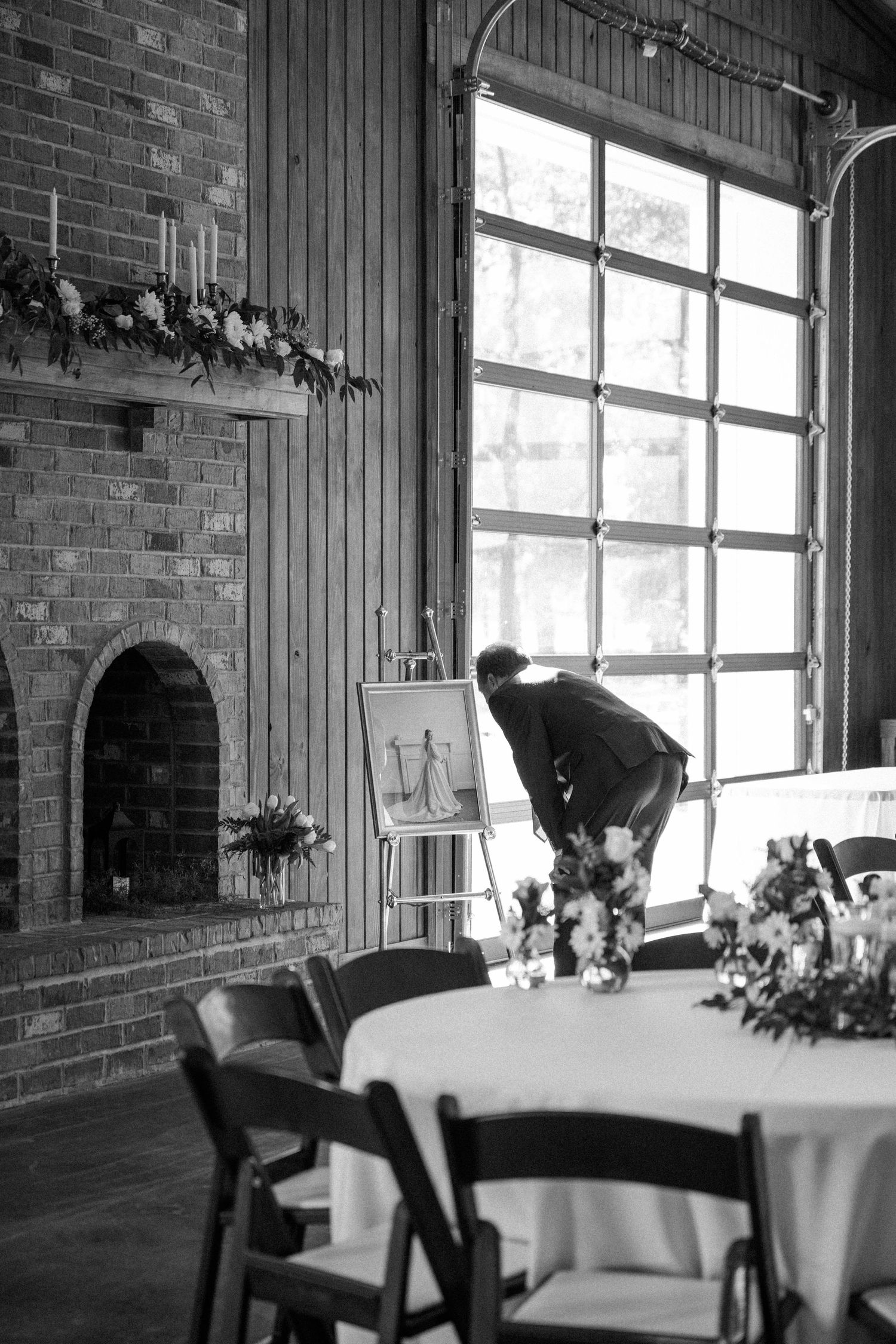 Black and white photo of a man arranging floral centerpieces on a round table inside a brick-fireplace wedding venue.