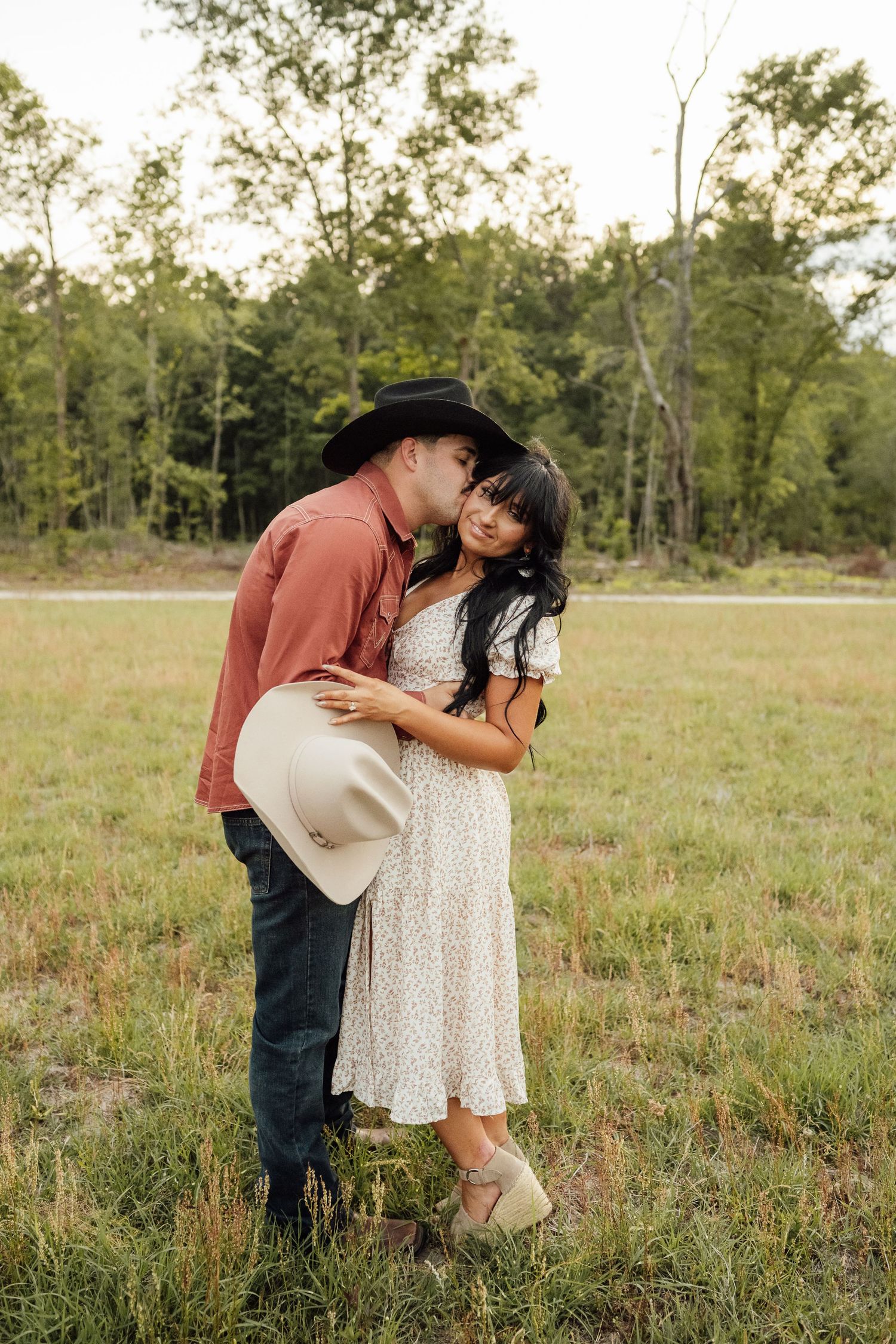 Man in a black cowboy hat kisses his fiancée on the forehead in a grassy field, holding a cream cowboy hat.
