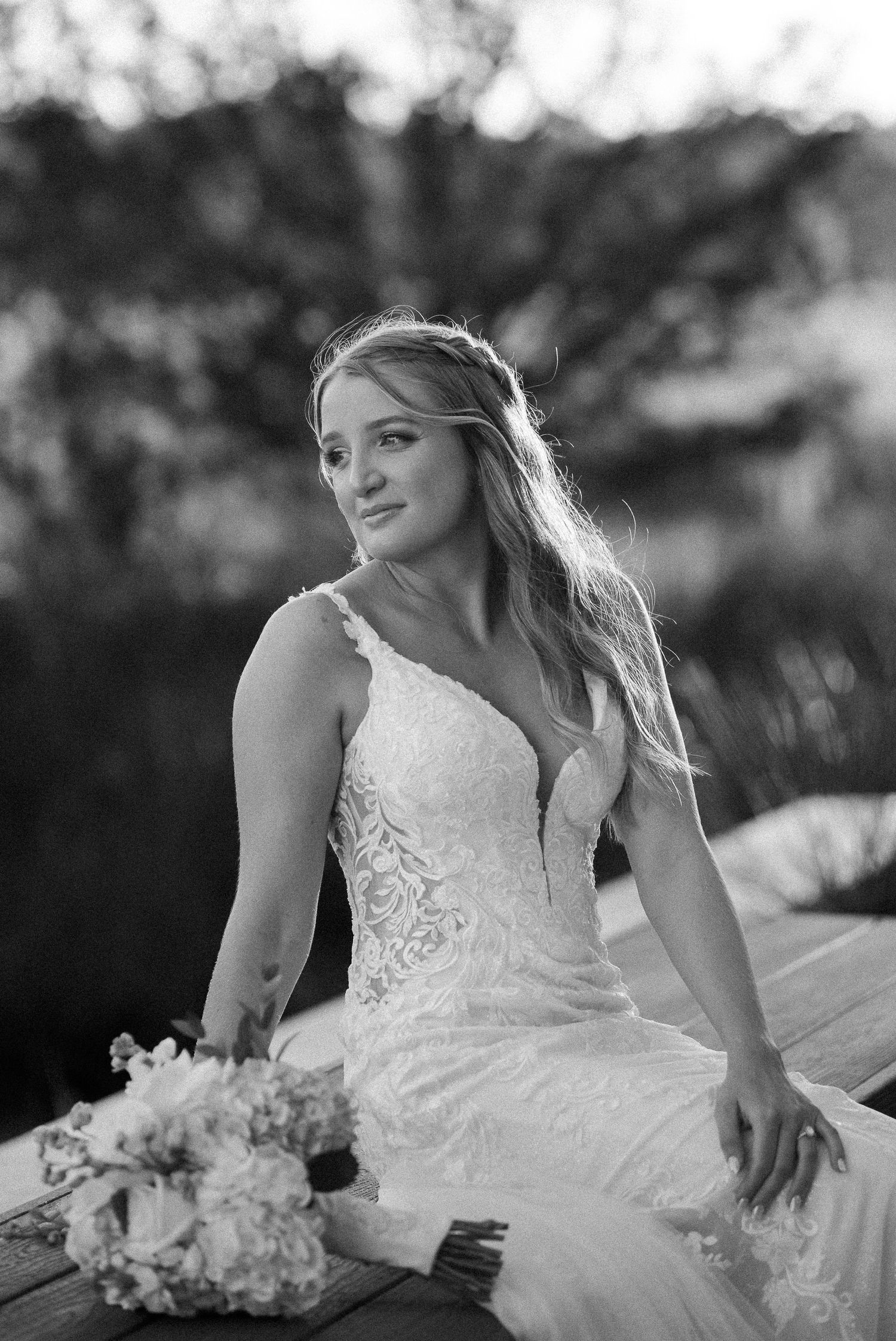 Black and white portrait of a bride in a lace gown with a bouquet, gazing away with her long hair flowing in soft light.