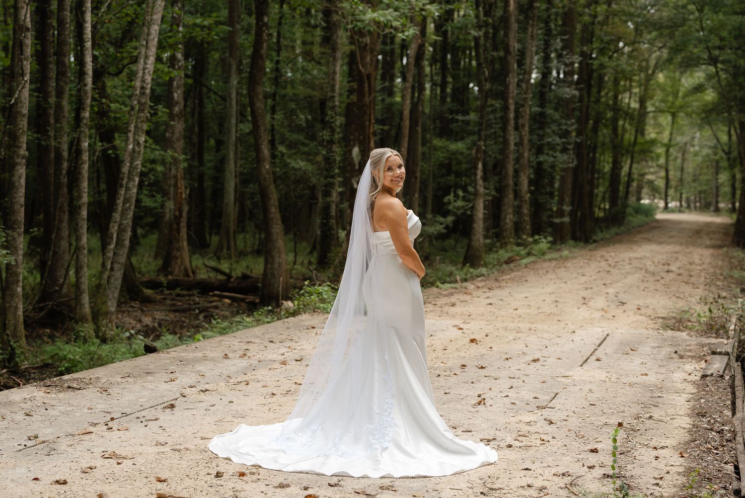 Bride in a long flowing wedding gown and veil stands on a sandy forest road lined with tall trees, glancing over her shoulder.
