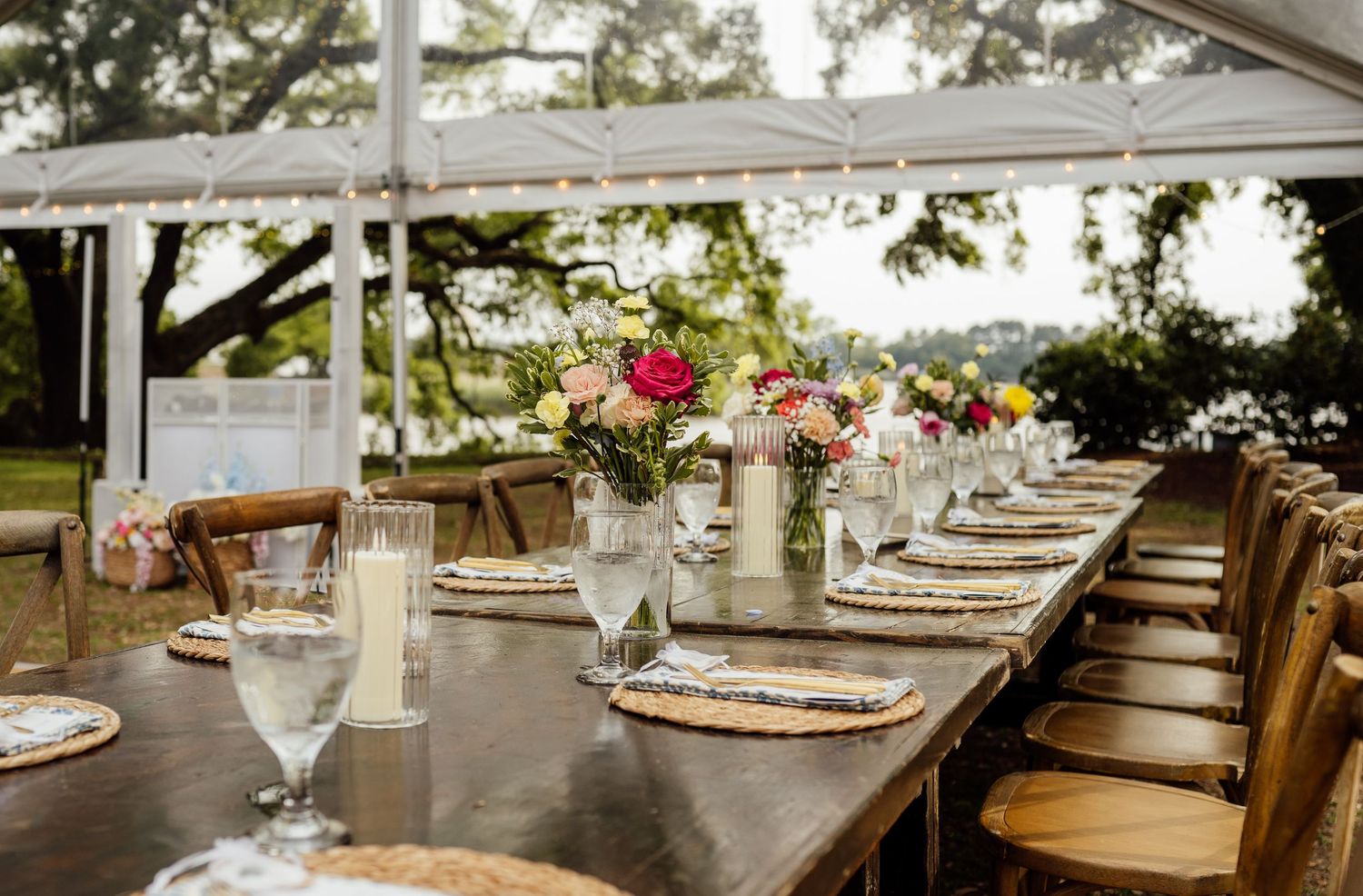 Long wooden reception table under a white tent set with woven chargers, glassware, candles and pink and peach floral centerpieces.