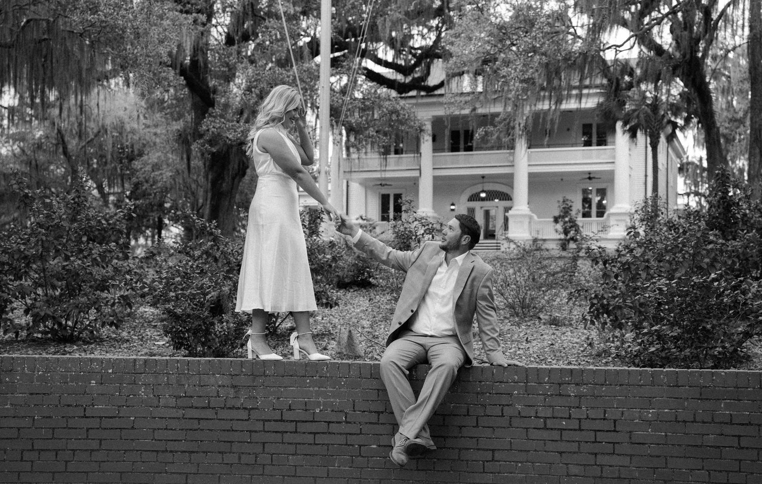 Black and white engagement photo of a woman holding hands with a seated man on a brick wall near a historic home.