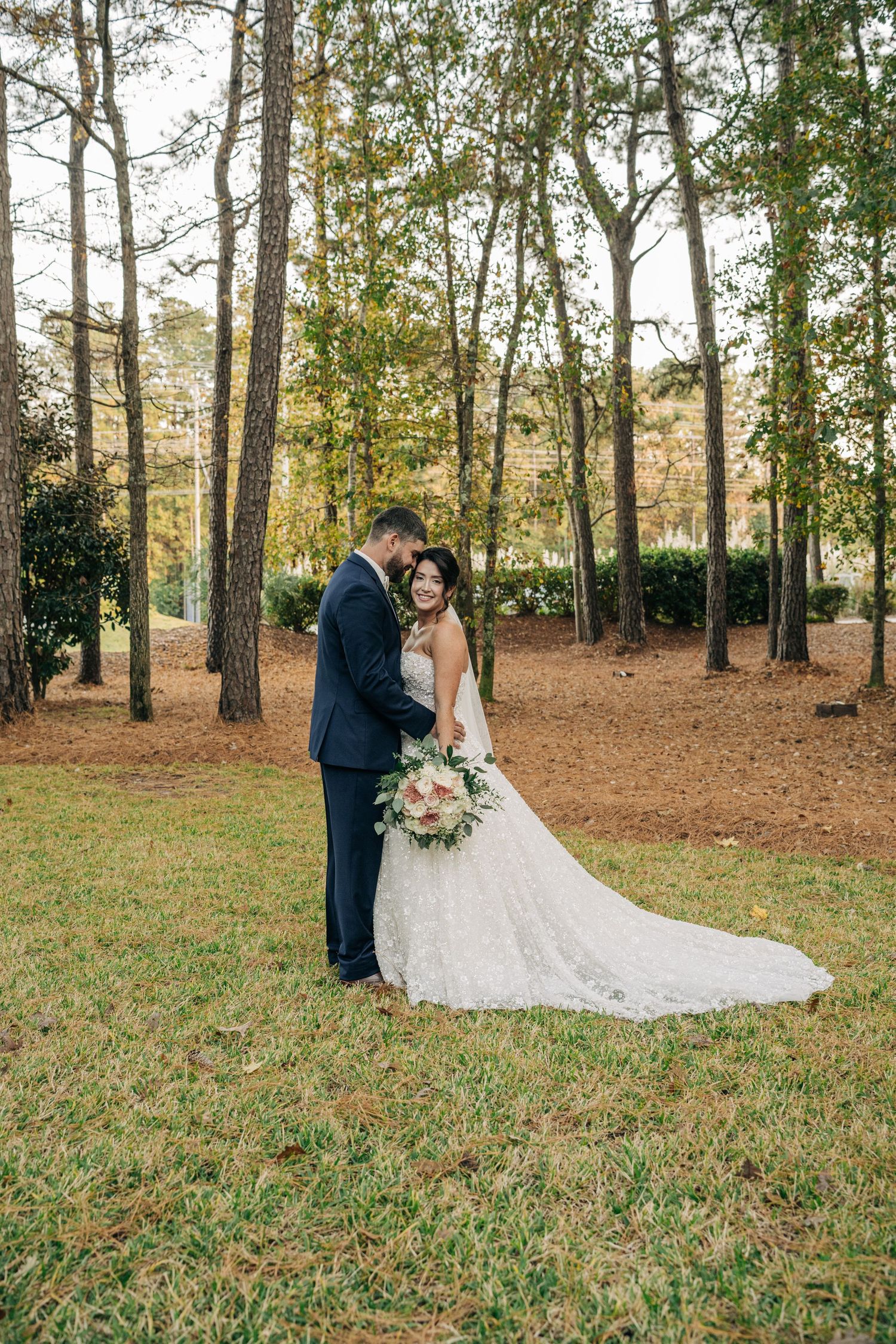 Bride in a lace wedding gown and groom in a navy suit embrace on a grassy lawn surrounded by tall pine trees.