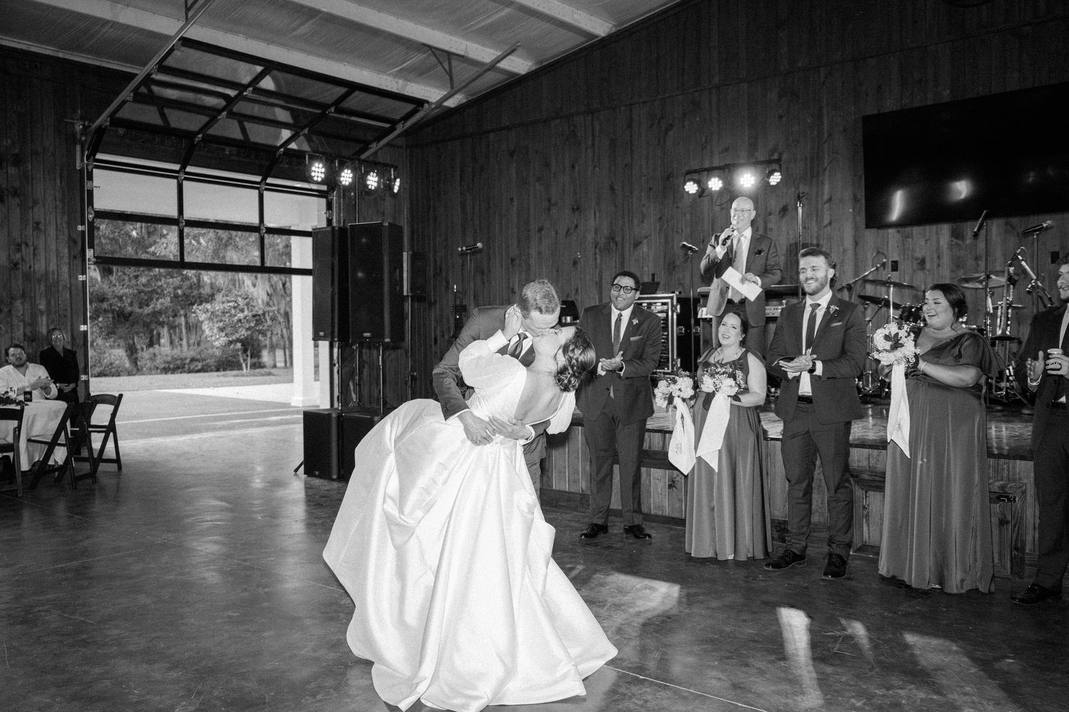 A groom dips his bride in a black and white wedding reception dance while a live band and guests watch in a rustic barn venue.