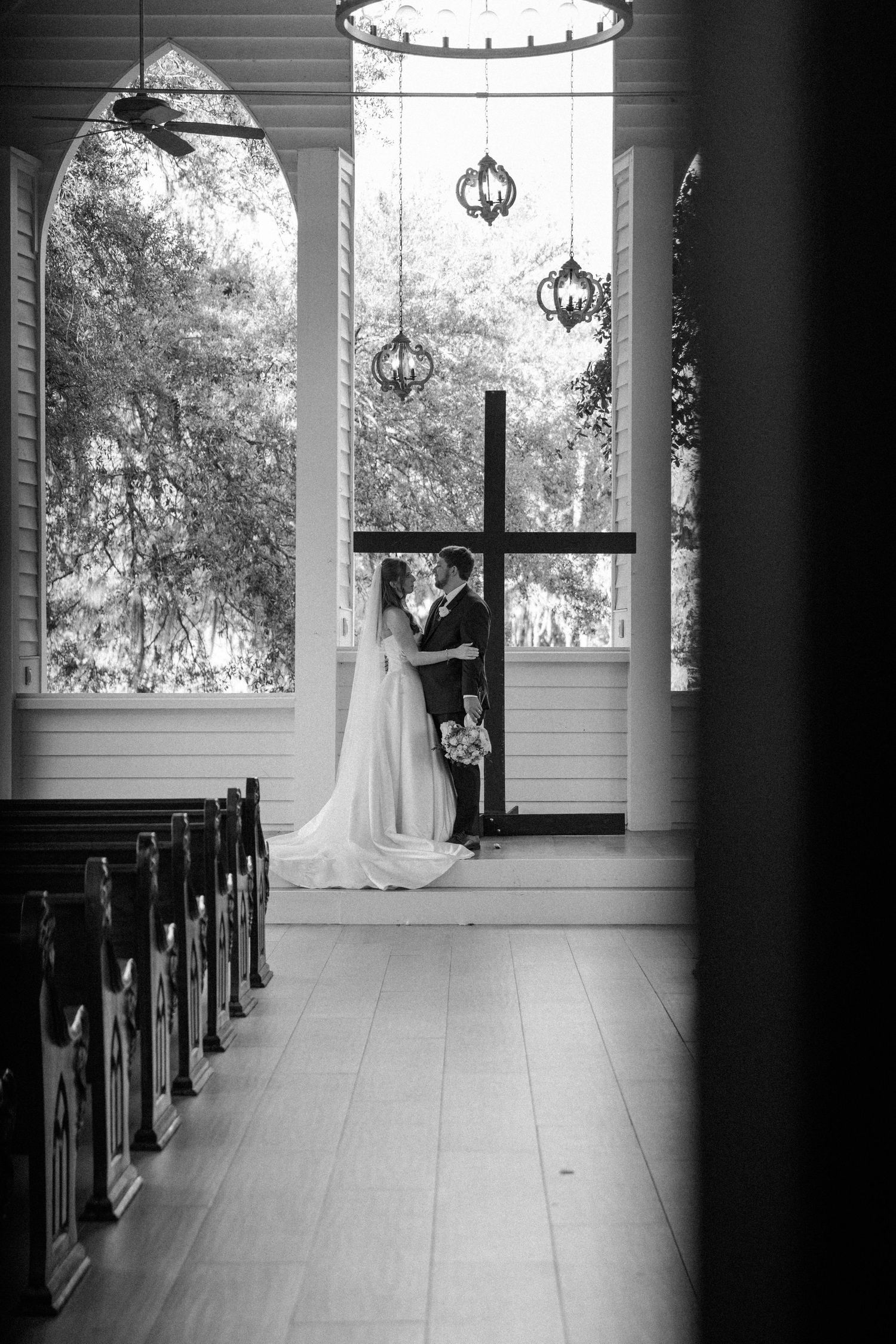 Bride and groom embrace at the front of an empty chapel before a large wooden cross, with pews lining the aisle in black and white.