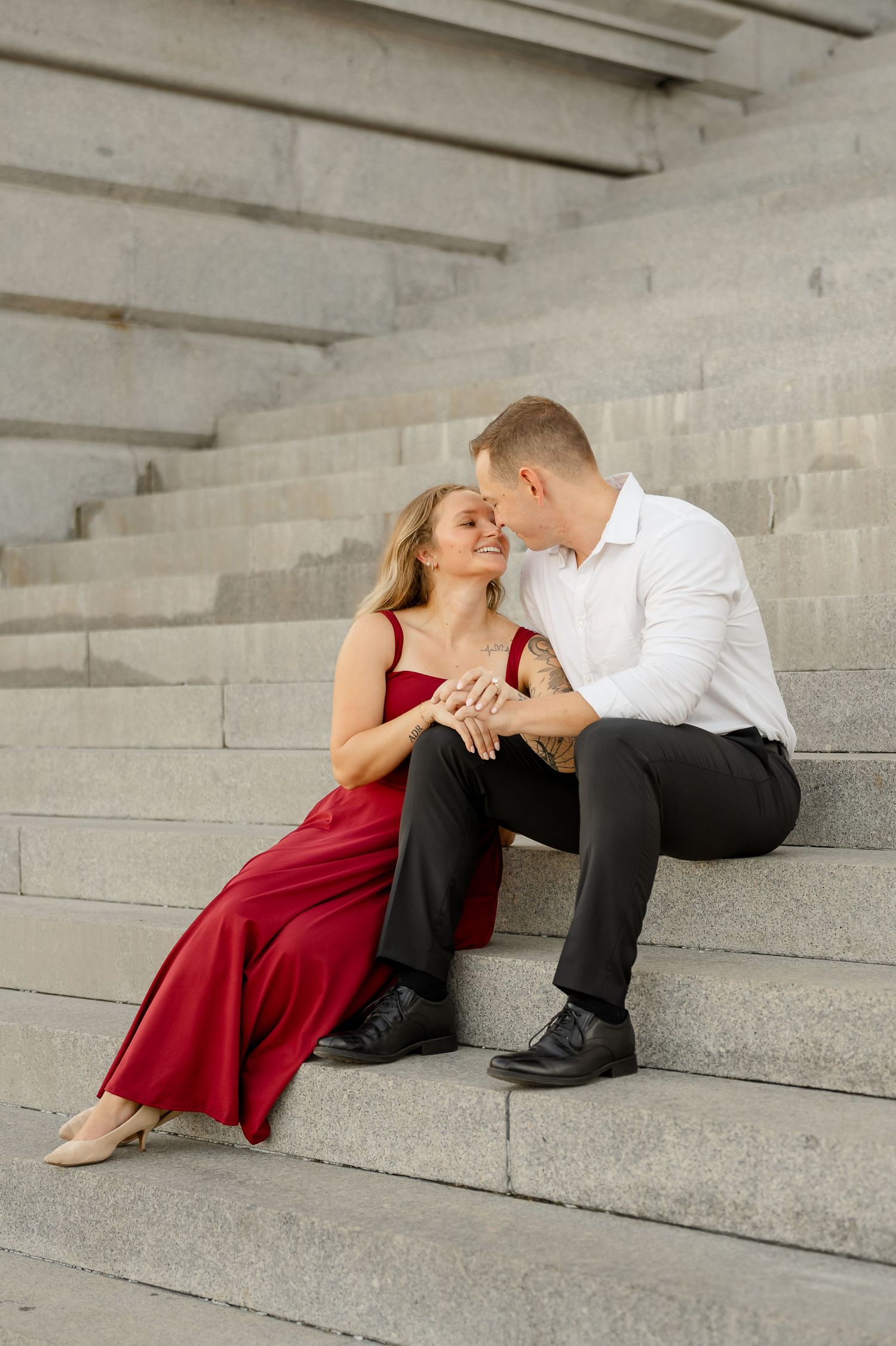 Couple sitting close on wide stone steps, the woman in a flowing red dress smiling as the man in a white shirt leans in.