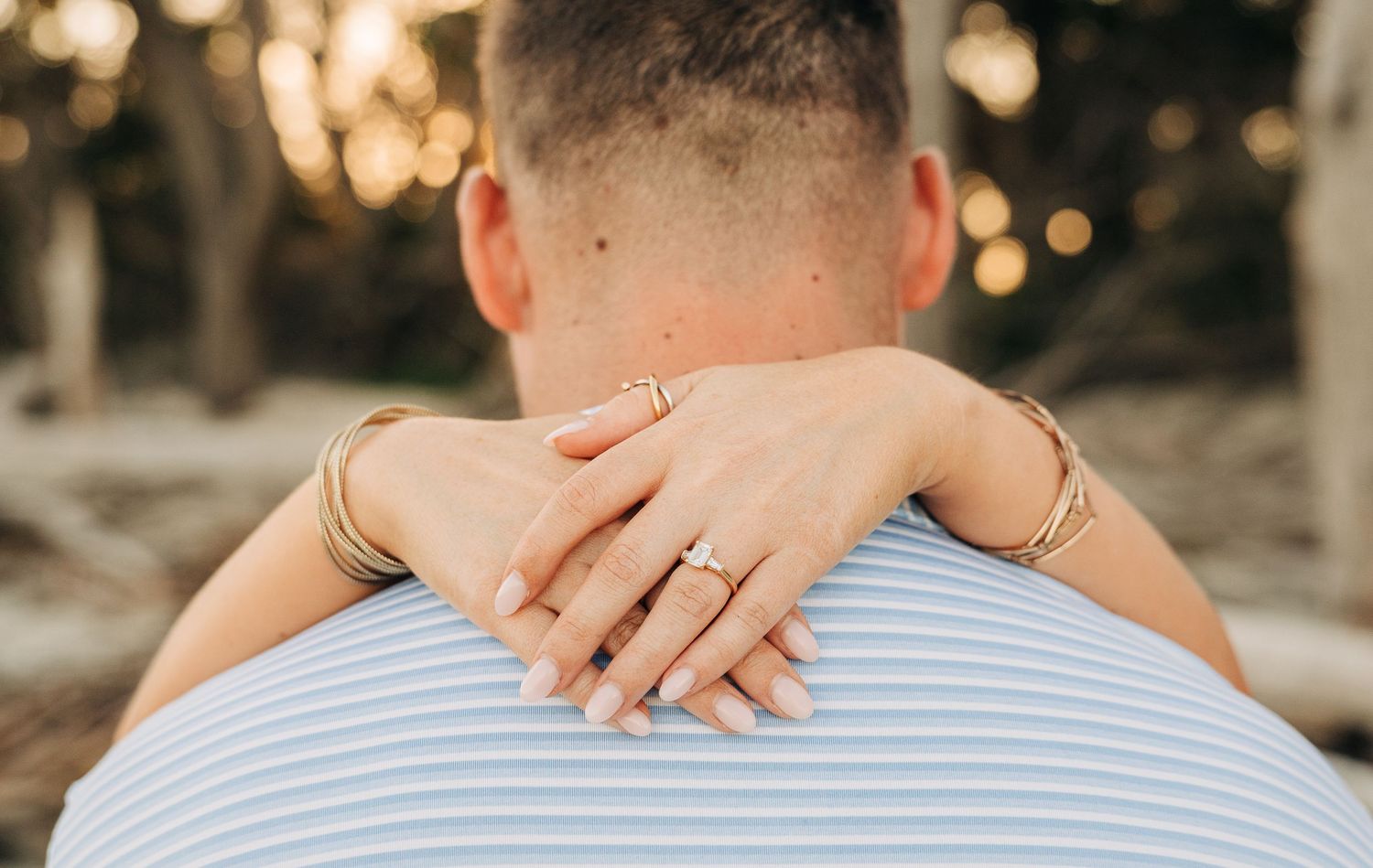 Close-up of a woman's hands with an engagement ring wrapped around a man's neck, with softly glowing lights in the background.
