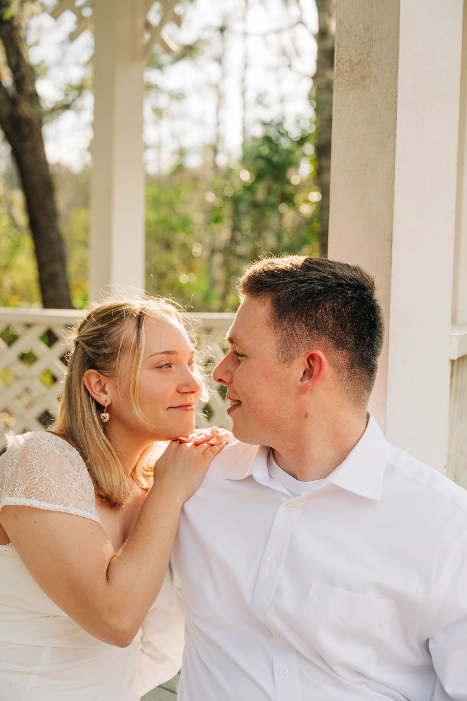 Smiling woman in a white lace dress rests her hands on a man's shoulder as they gaze at each other in a gazebo.