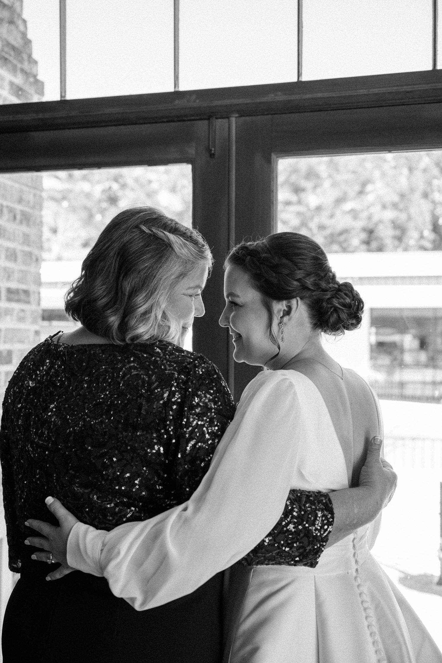 Black and white photo of a bride embracing her mother in a sparkly top as they share a tender moment by a window.