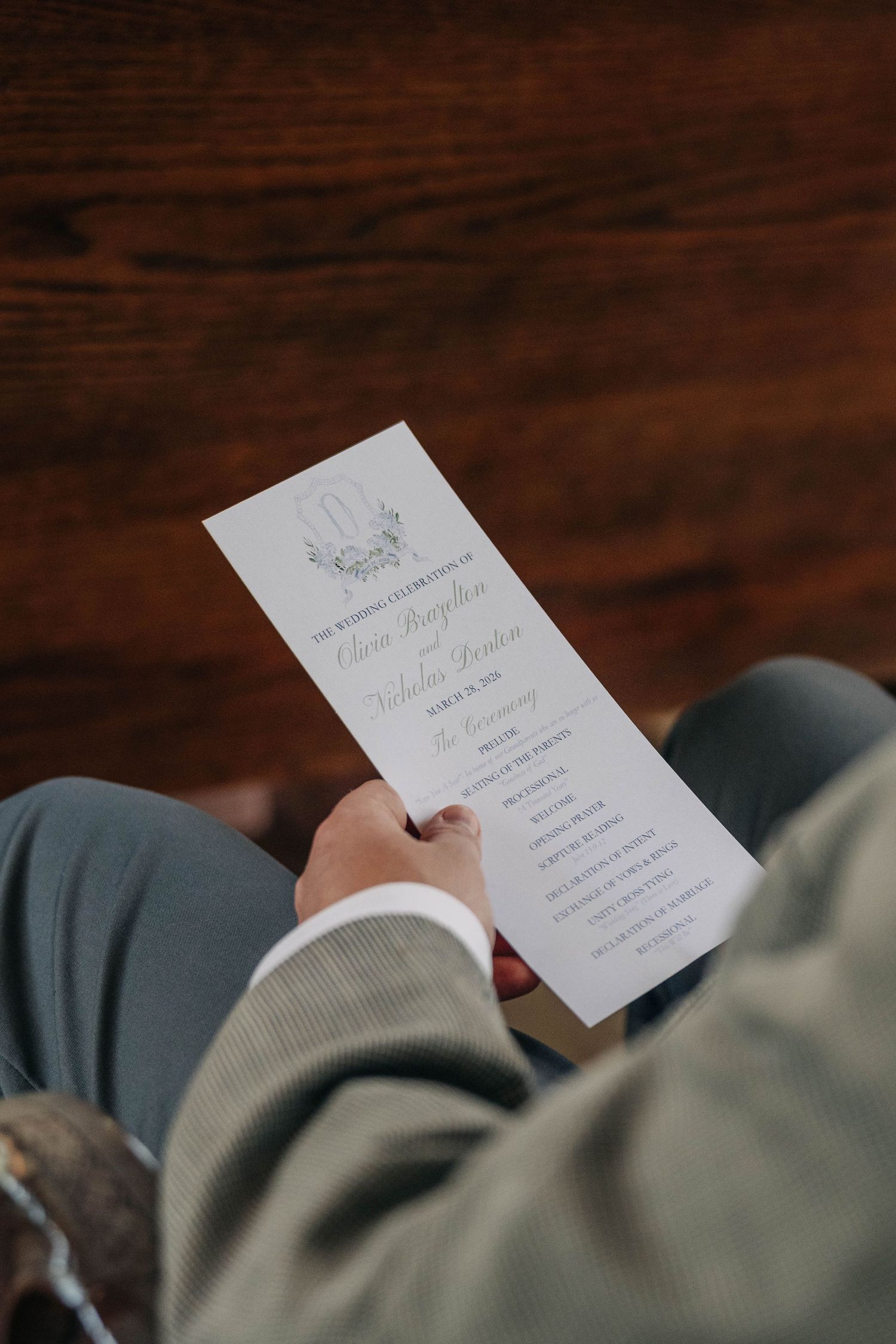 Wedding guest in a gray suit holds a printed ceremony program while seated on a wooden pew.